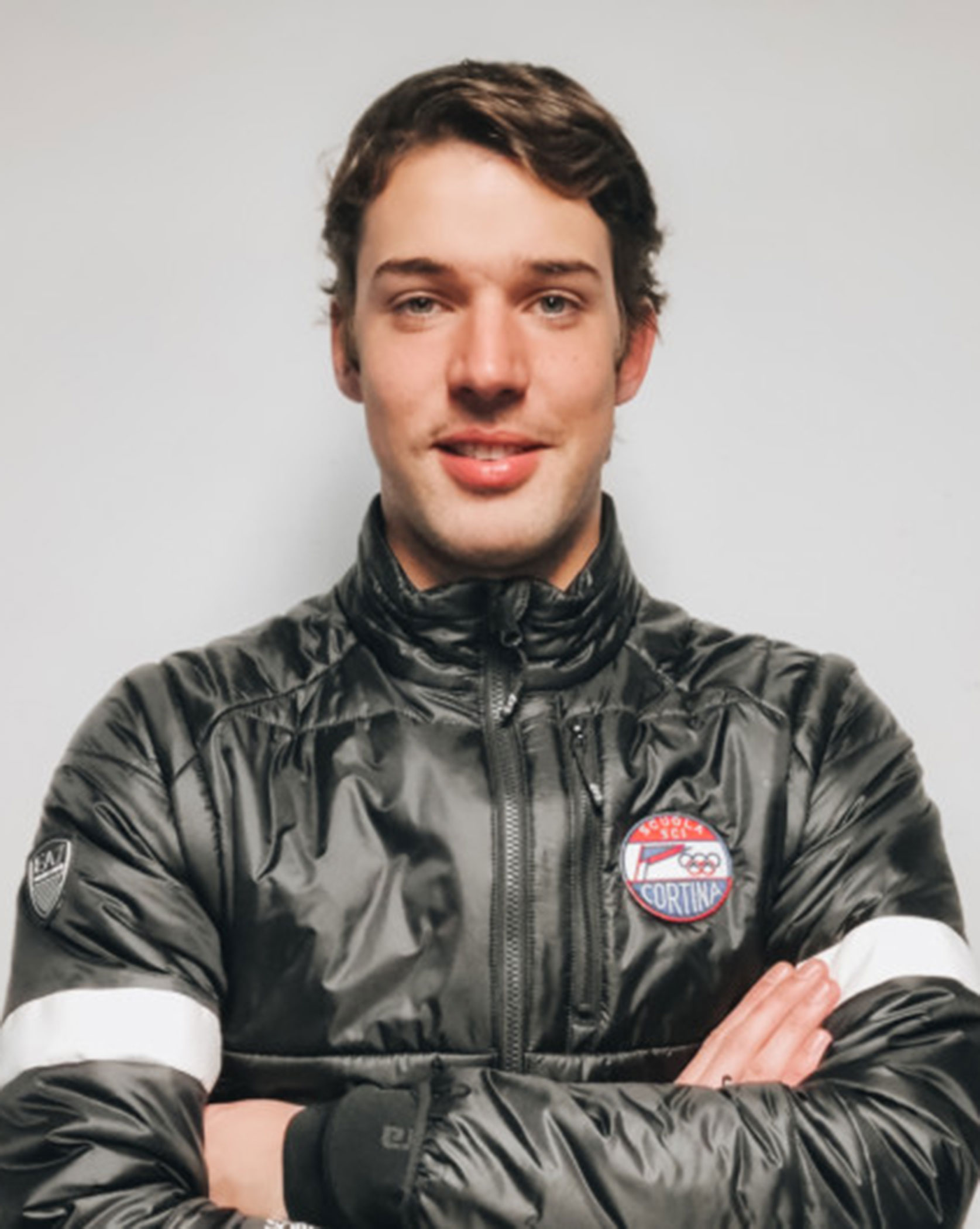 Man in a black puffer jacket with arms crossed, standing against a plain light-colored background. - Scuola Sci Cortina