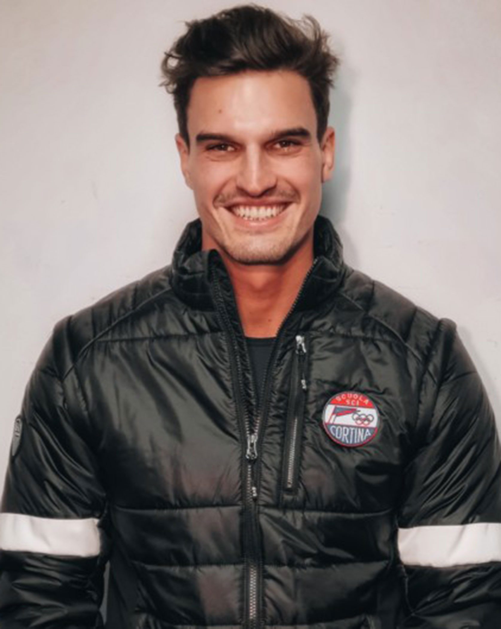Man in a black puffer jacket with a Cortina patch, smiling in front of a plain light background. - Scuola Sci Cortina