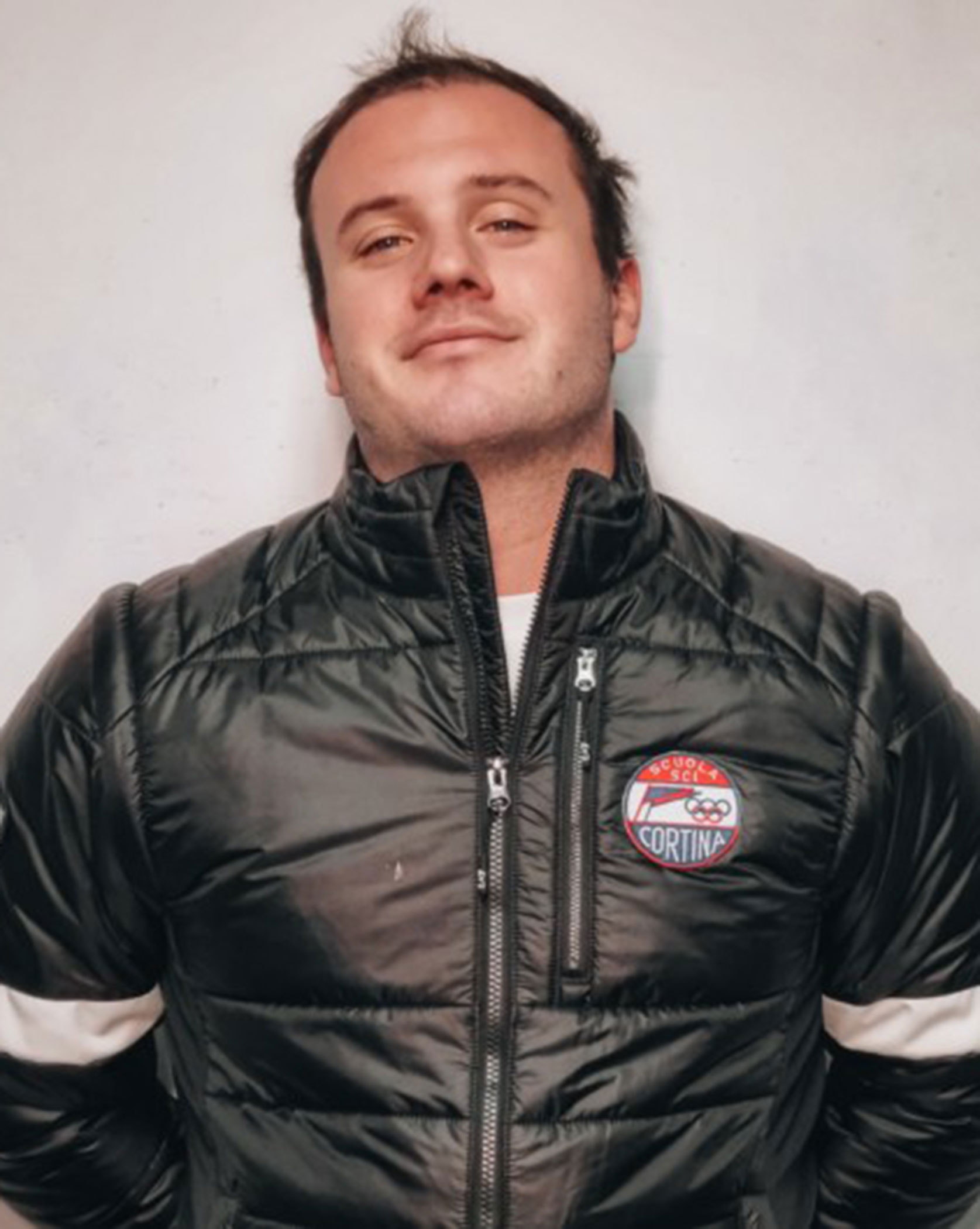 Man in a black puffer jacket with a Cortina patch stands in front of a plain light-colored wall. - Scuola Sci Cortina