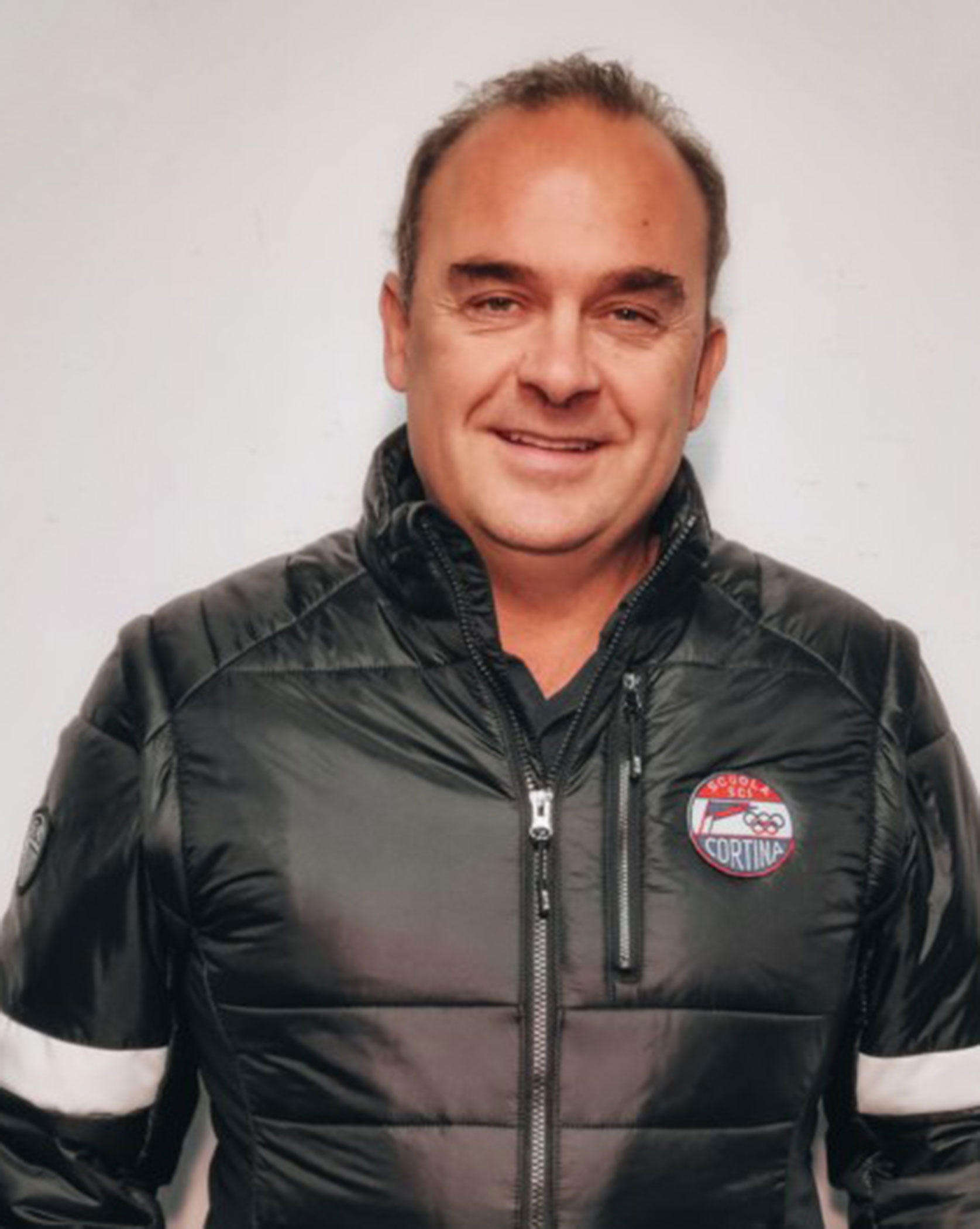 Man in a black puffer jacket with a Cortina logo, standing against a plain light background, smiling. - Scuola Sci Cortina