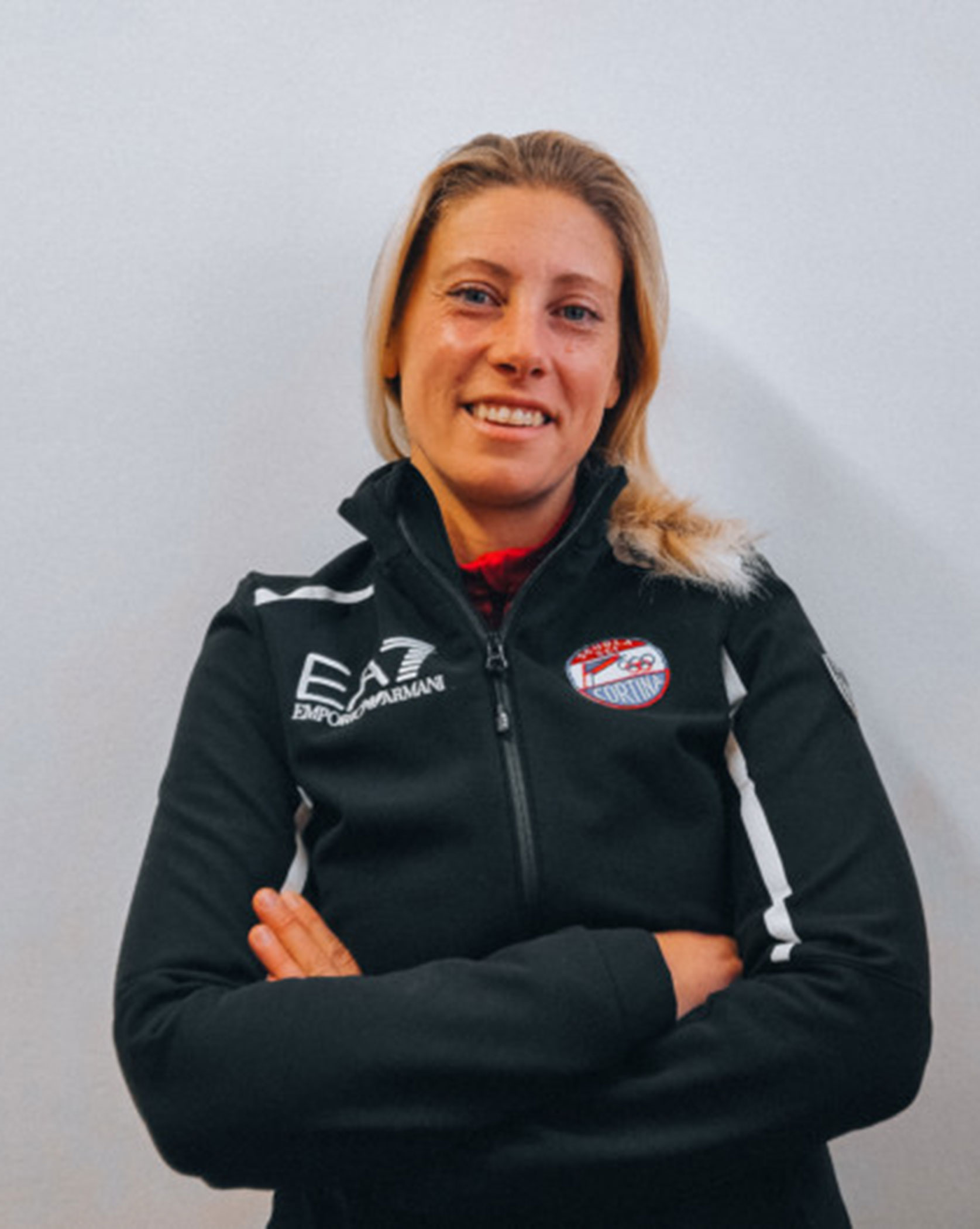 Woman in a black sports jacket with arms crossed, smiling, standing against a plain white background. - Scuola Sci Cortina