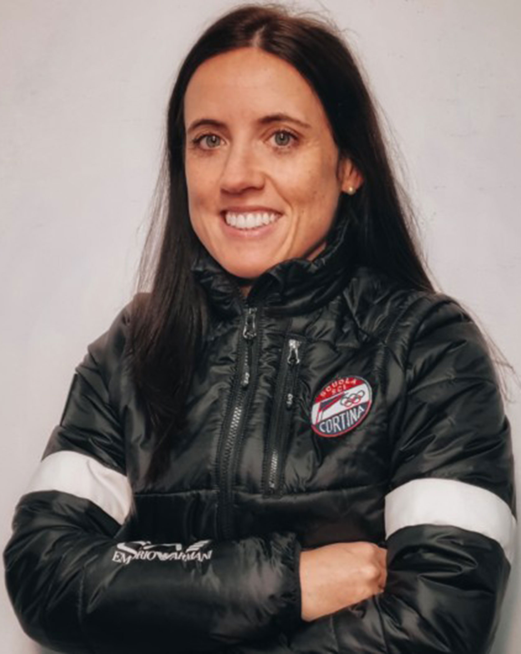 Woman with long dark hair in a black jacket, arms crossed, smiling at the camera against a plain background. - Scuola Sci Cortina