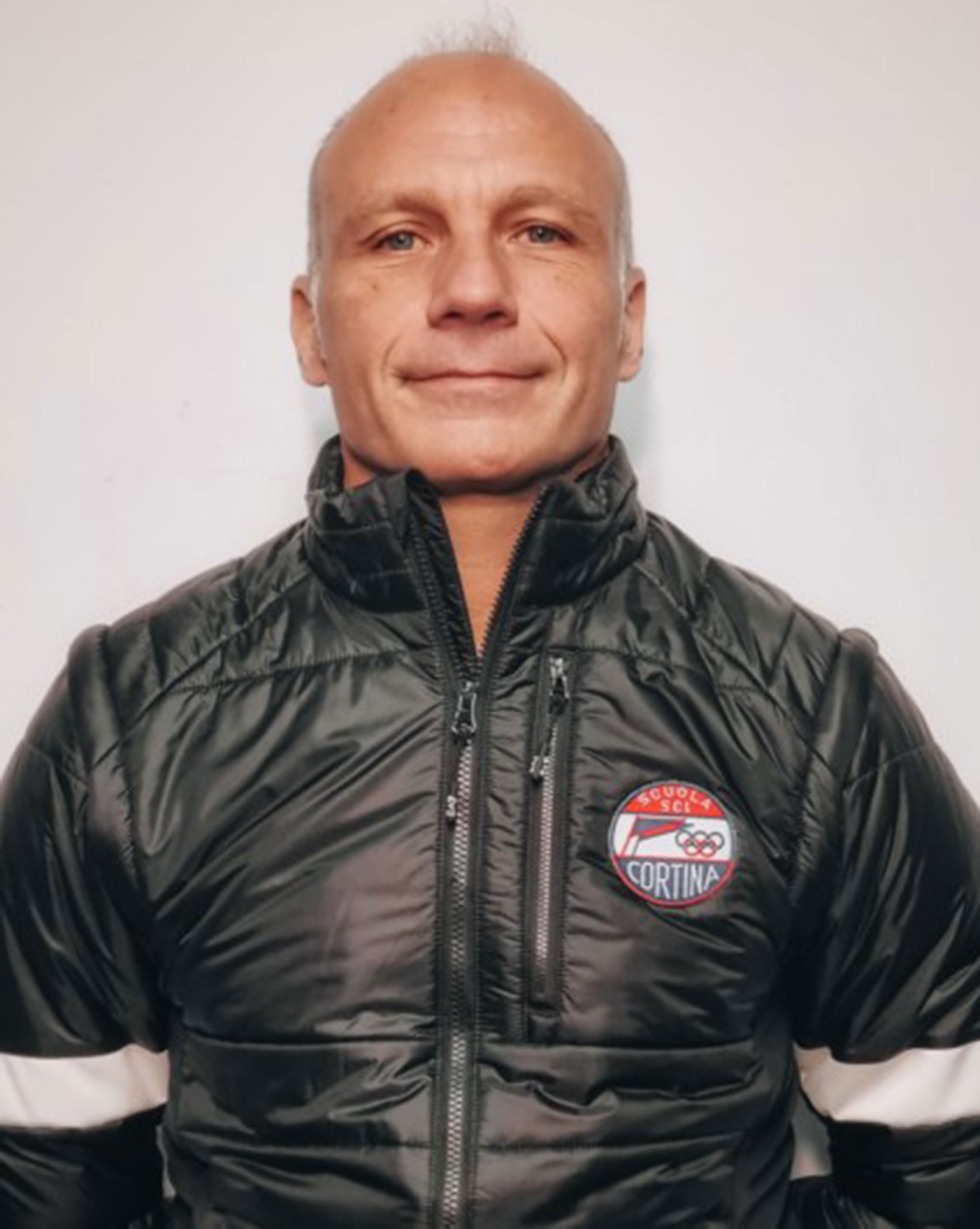 Bald man in a black Cortina jacket stands against a plain white background, looking at the camera. - Scuola Sci Cortina