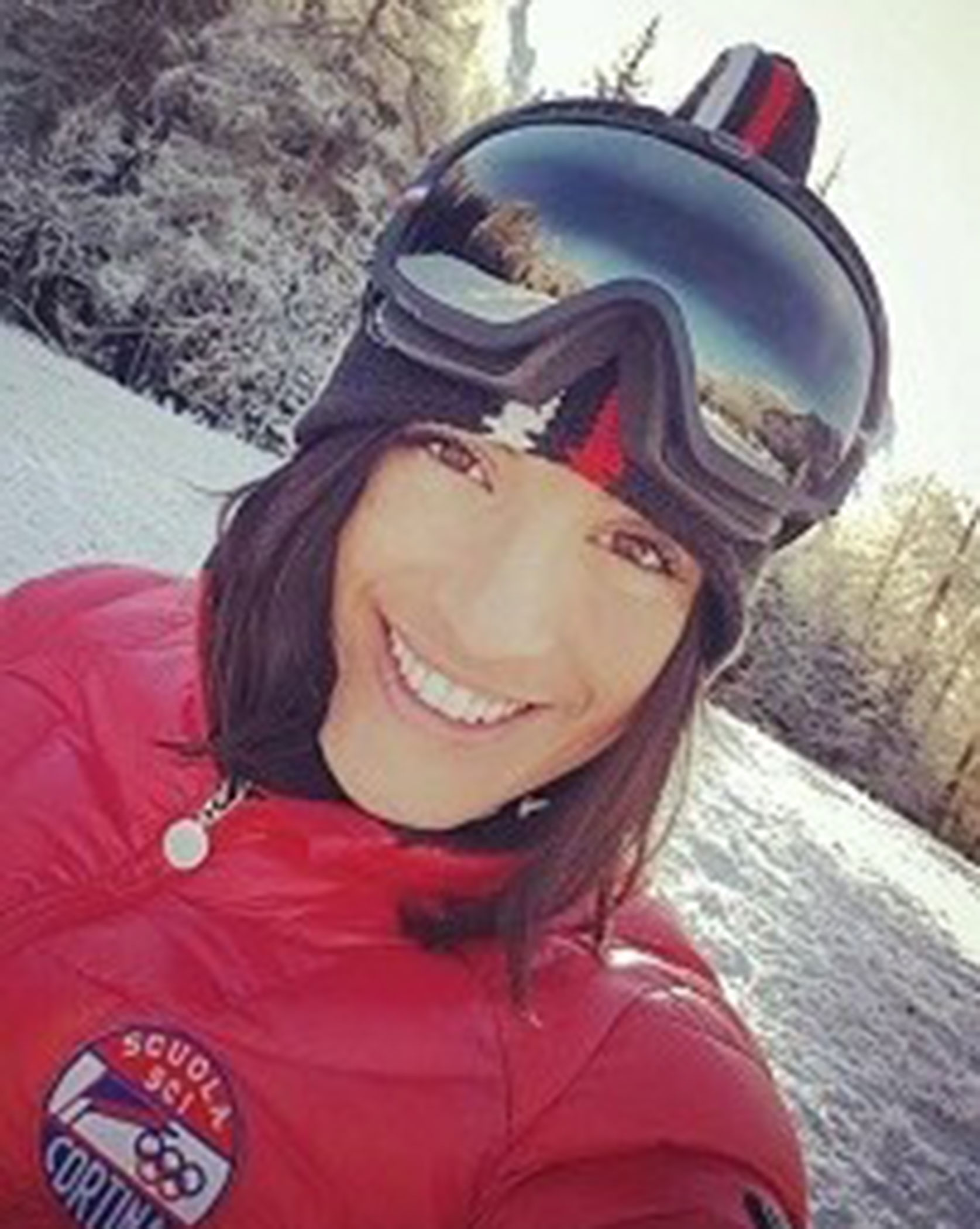 Woman in red ski jacket and goggles smiling outdoors in a snowy landscape, trees in the background. - Scuola Sci Cortina