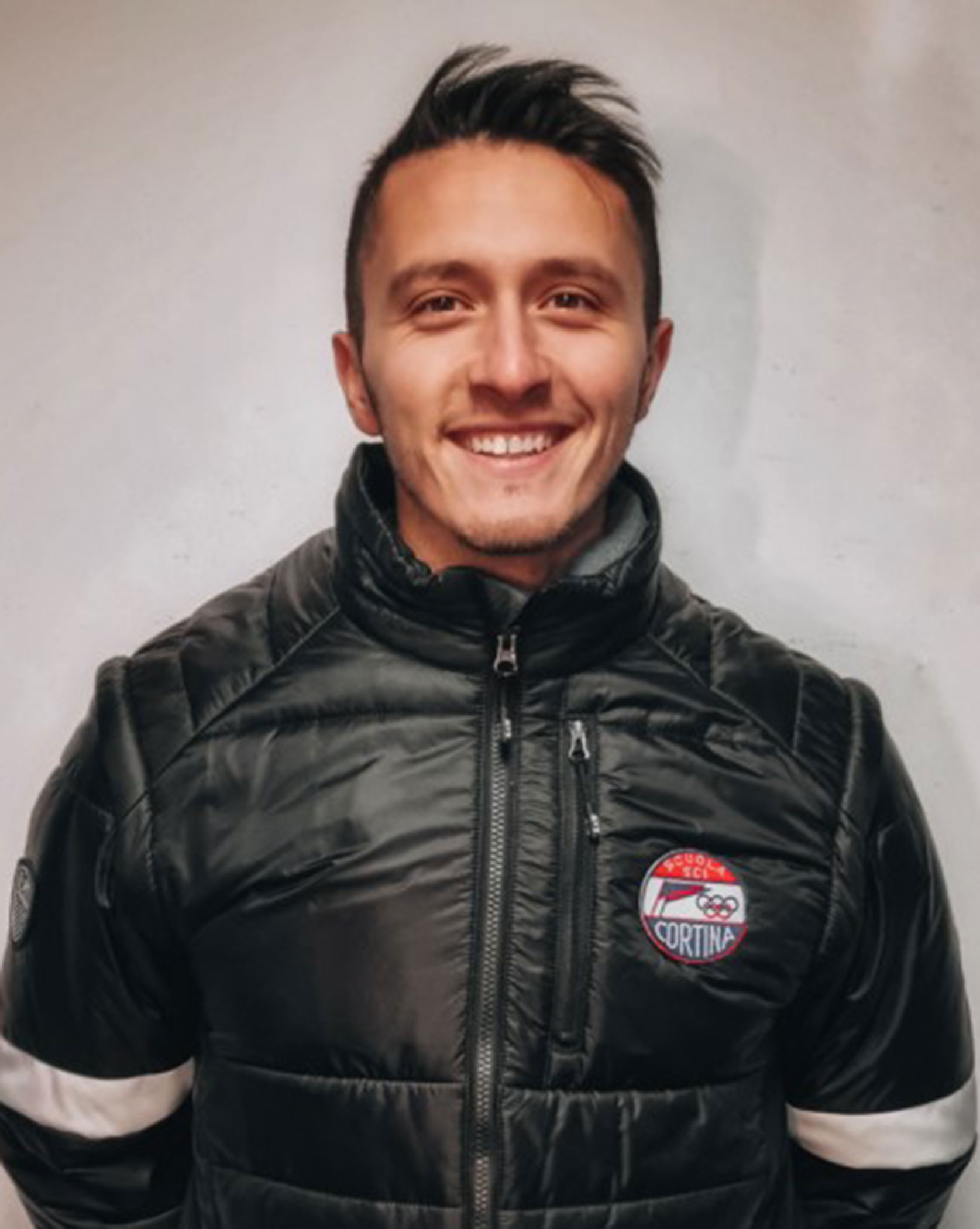 Smiling person wearing a black jacket with a Cortina patch, standing against a plain background. - Scuola Sci Cortina
