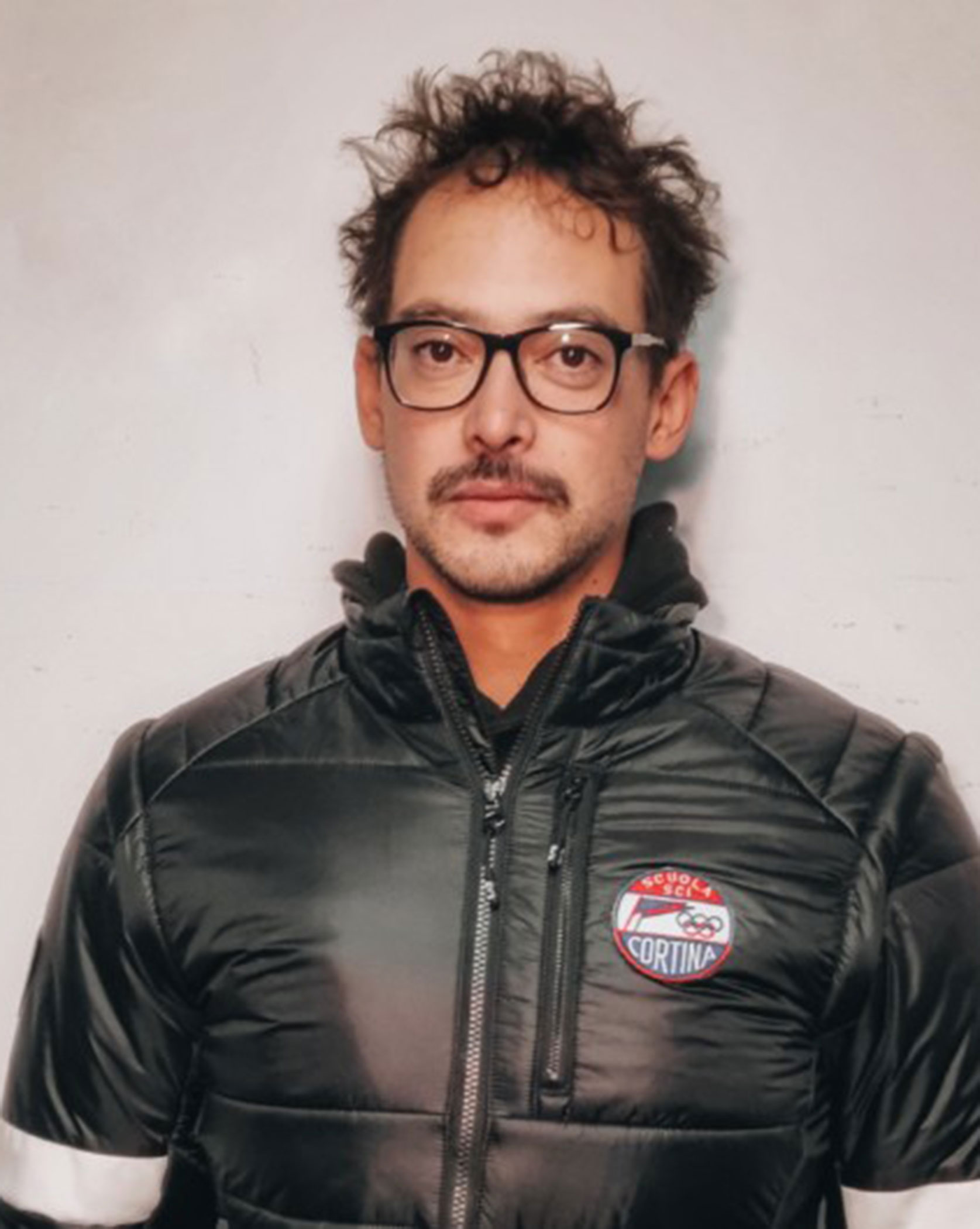 Man with glasses and mustache wearing a black jacket with a Cortina patch, standing against a white wall. - Scuola Sci Cortina