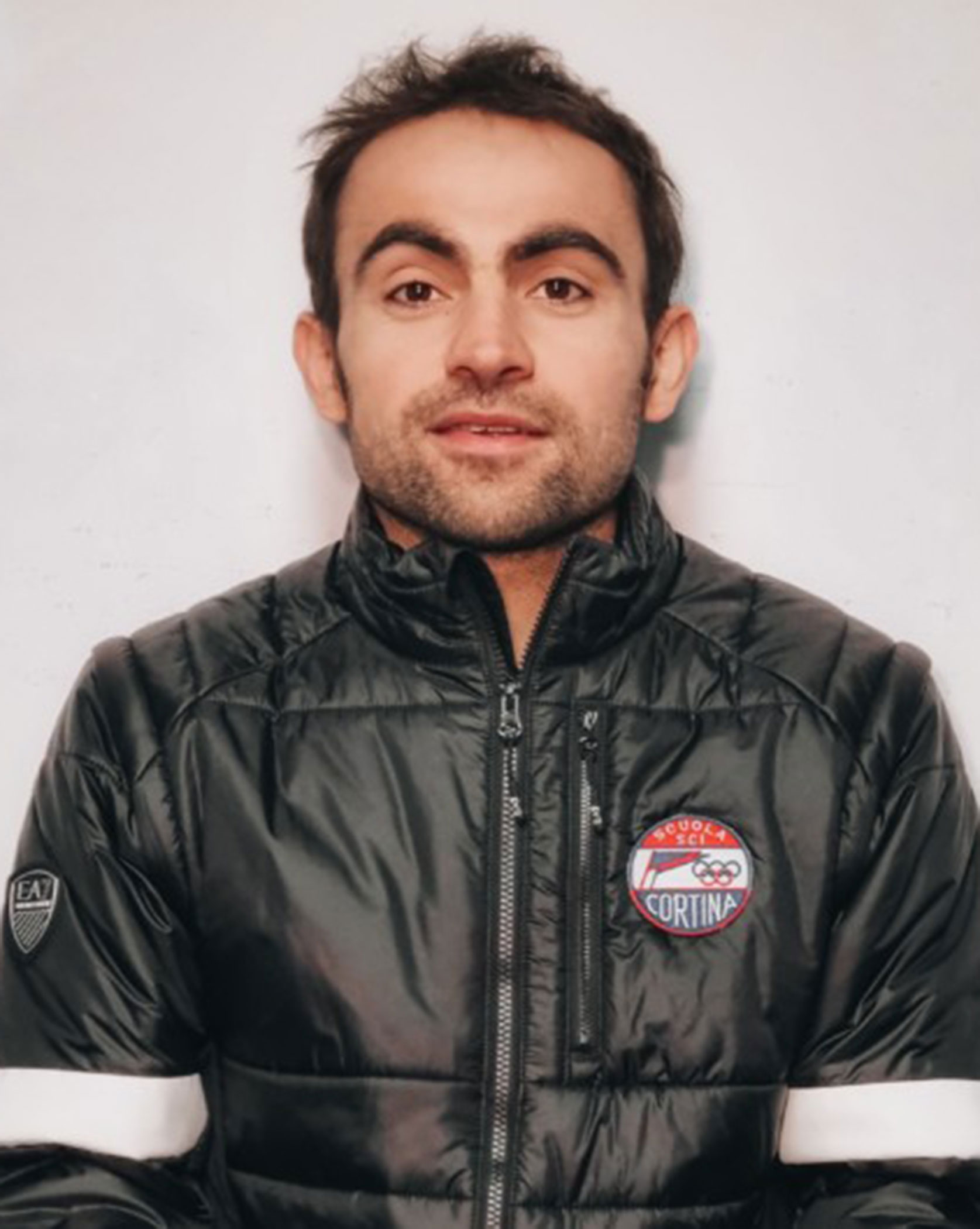 Man in a black puffer jacket with Cortina badge, standing against a plain light background. - Scuola Sci Cortina