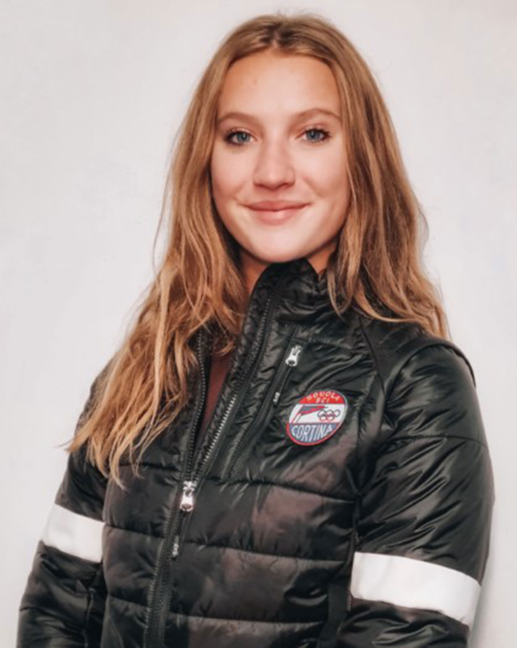 Young woman with long brown hair wearing a black puffer jacket with a Cortina patch, smiling. - Scuola Sci Cortina
