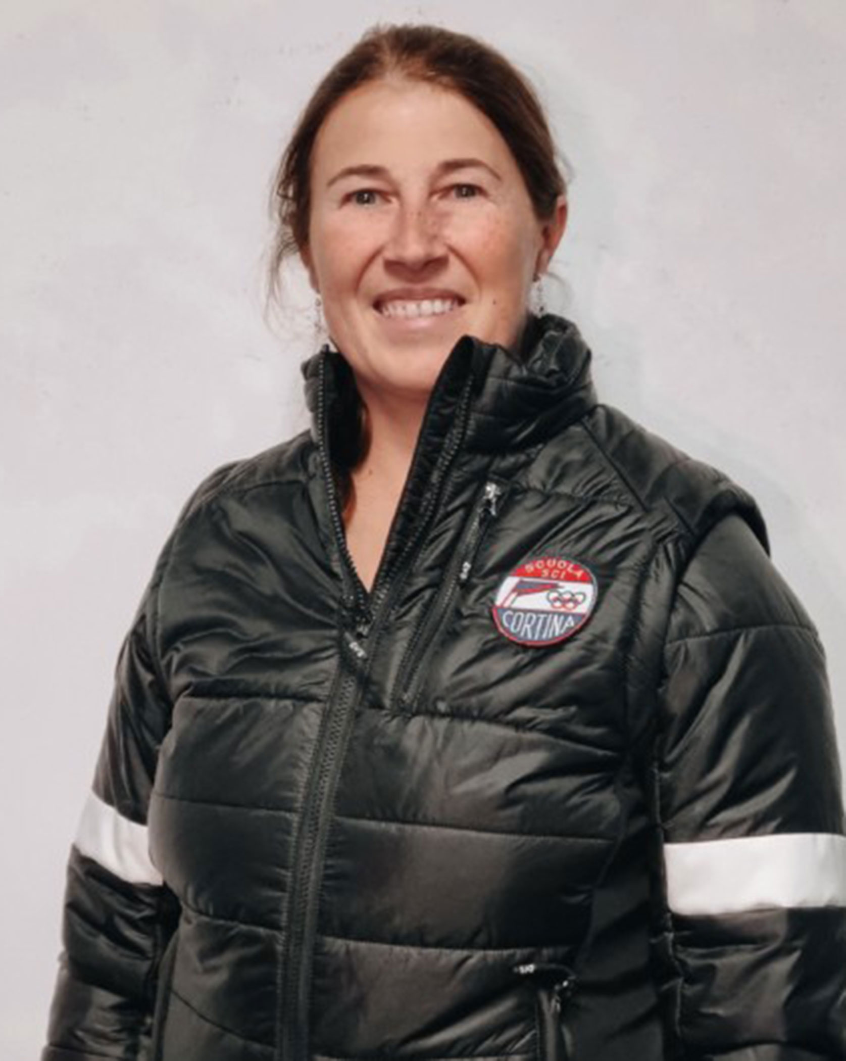Woman in a black puffer jacket with a Cortina patch, smiling in front of a plain light background. - Scuola Sci Cortina