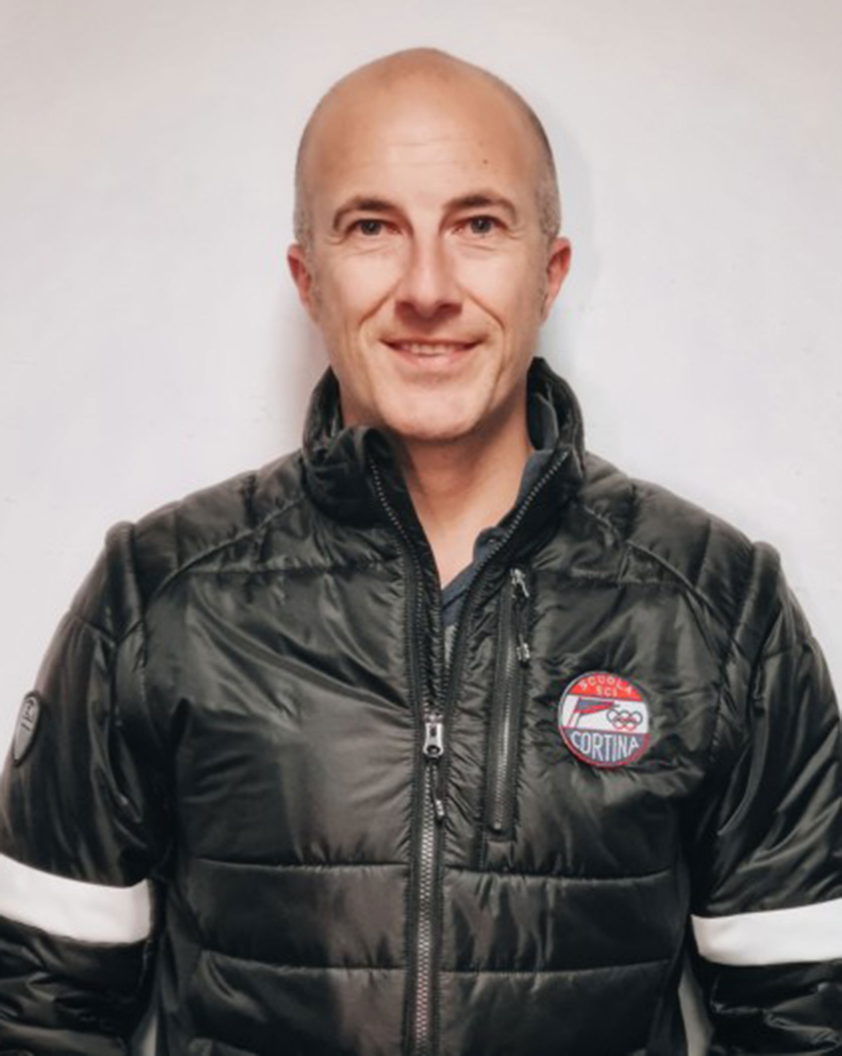Smiling man in a black jacket with a Cortina logo, standing against a plain white background. - Scuola Sci Cortina