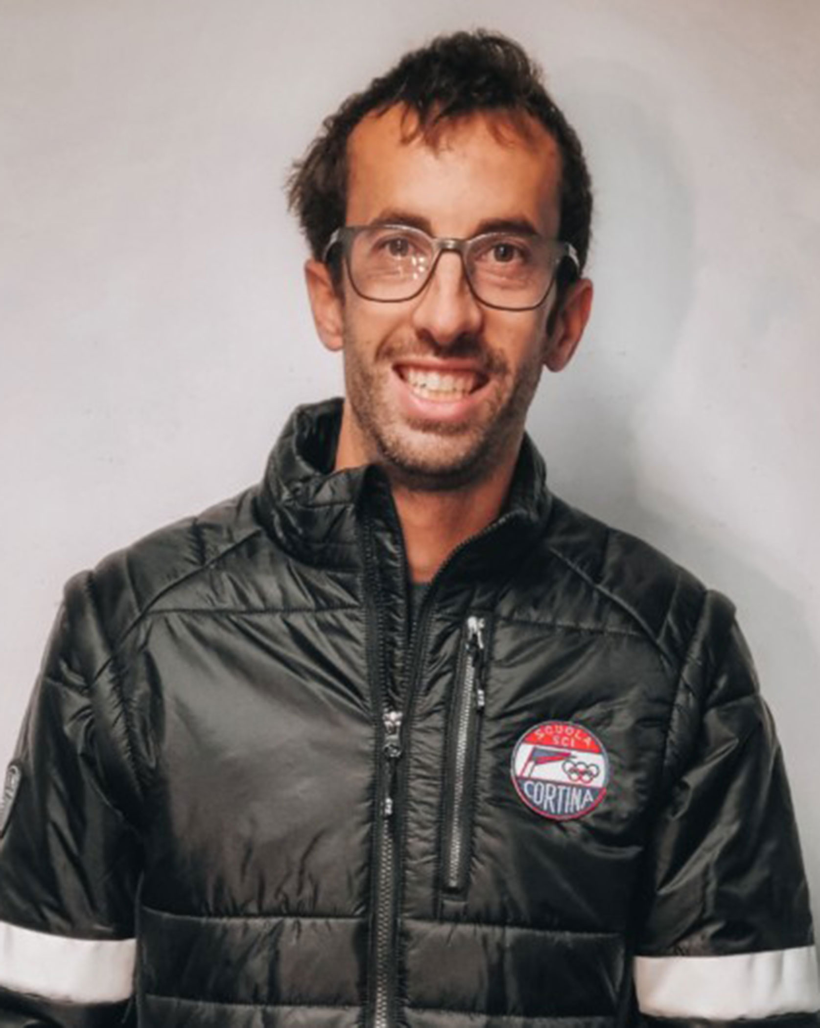 Man with glasses, wearing a black jacket with a Cortina logo, smiles at the camera against a plain background. - Scuola Sci Cortina