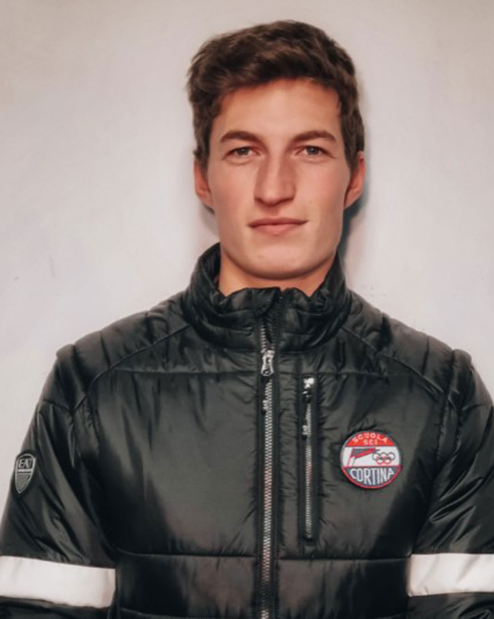 Young man in a black puffer jacket with a Cortina logo, standing against a plain light background. - Scuola Sci Cortina