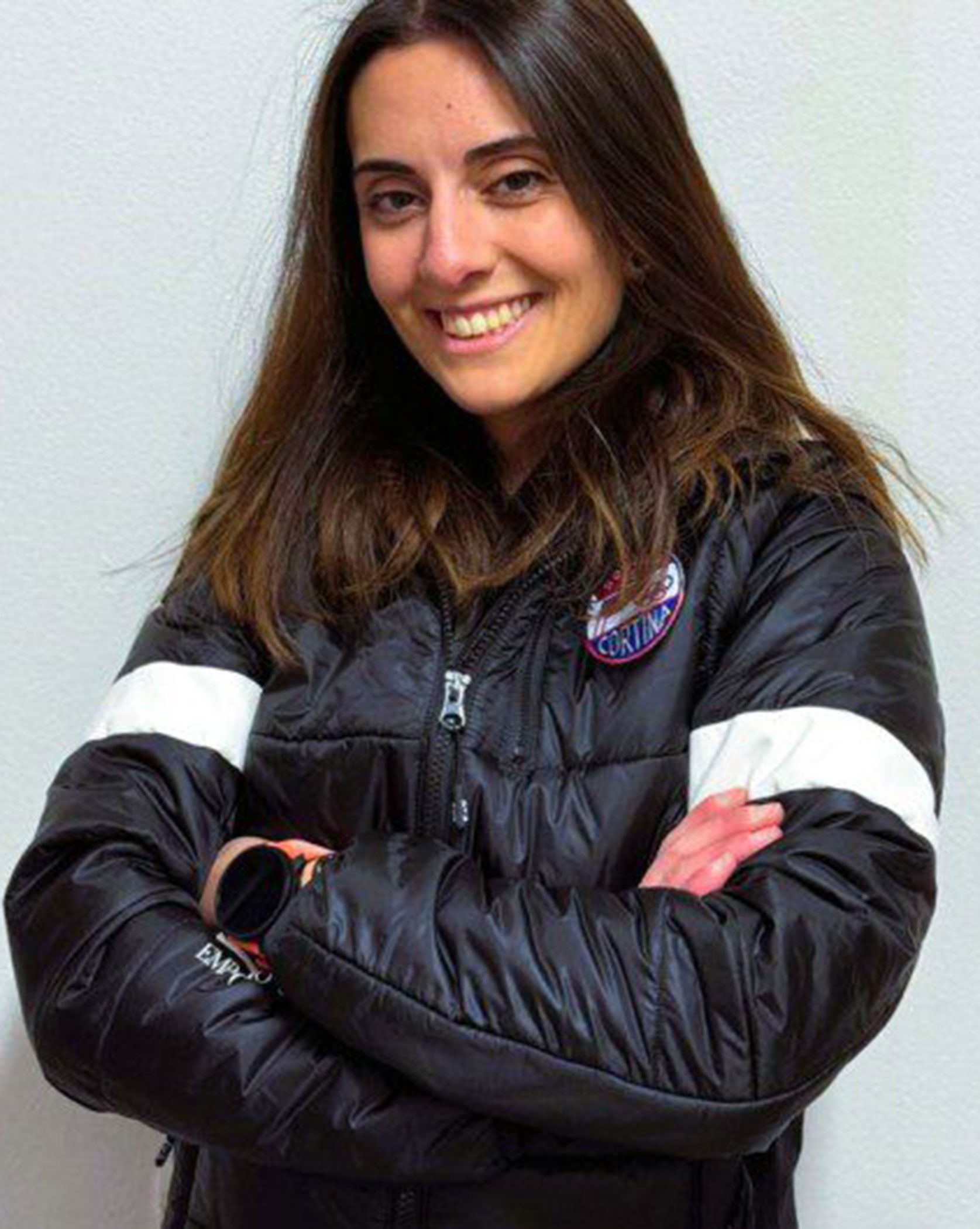 Woman in a black puffer jacket with arms crossed, smiling, standing against a plain white wall. - Scuola Sci Cortina