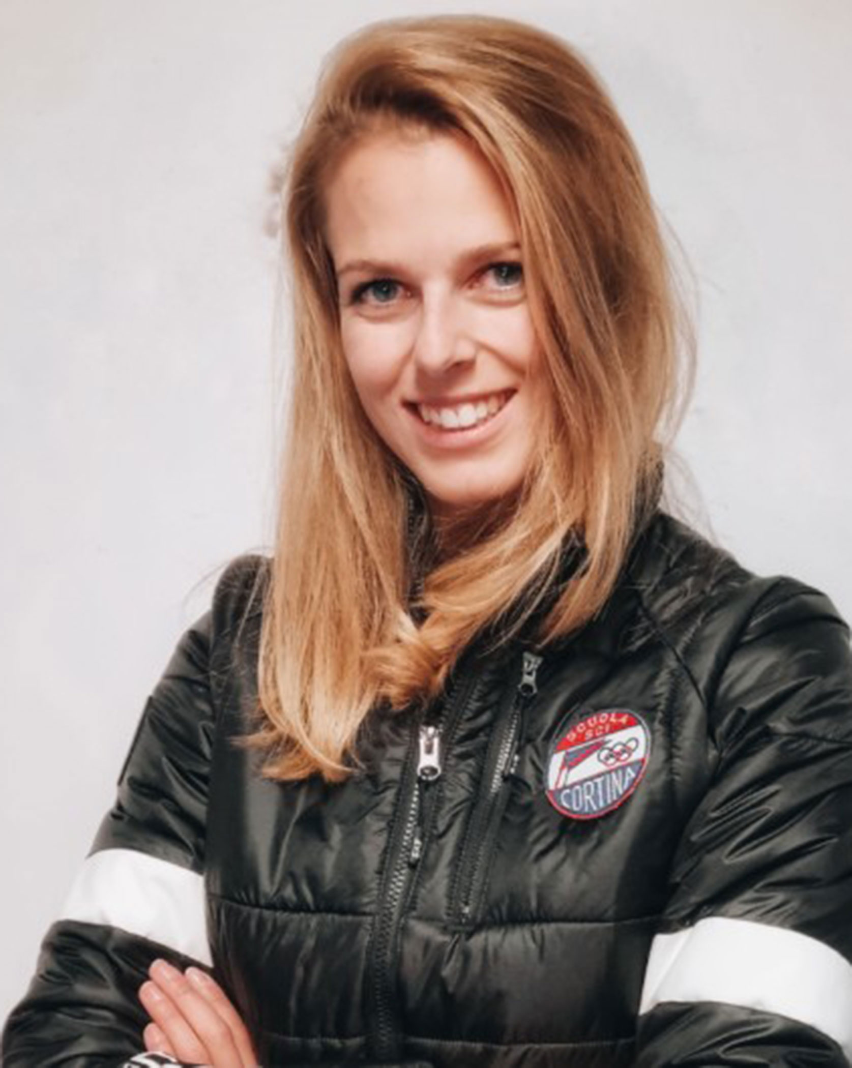 Smiling woman with long blonde hair wearing a black jacket with a Cortina patch, arms crossed. - Scuola Sci Cortina
