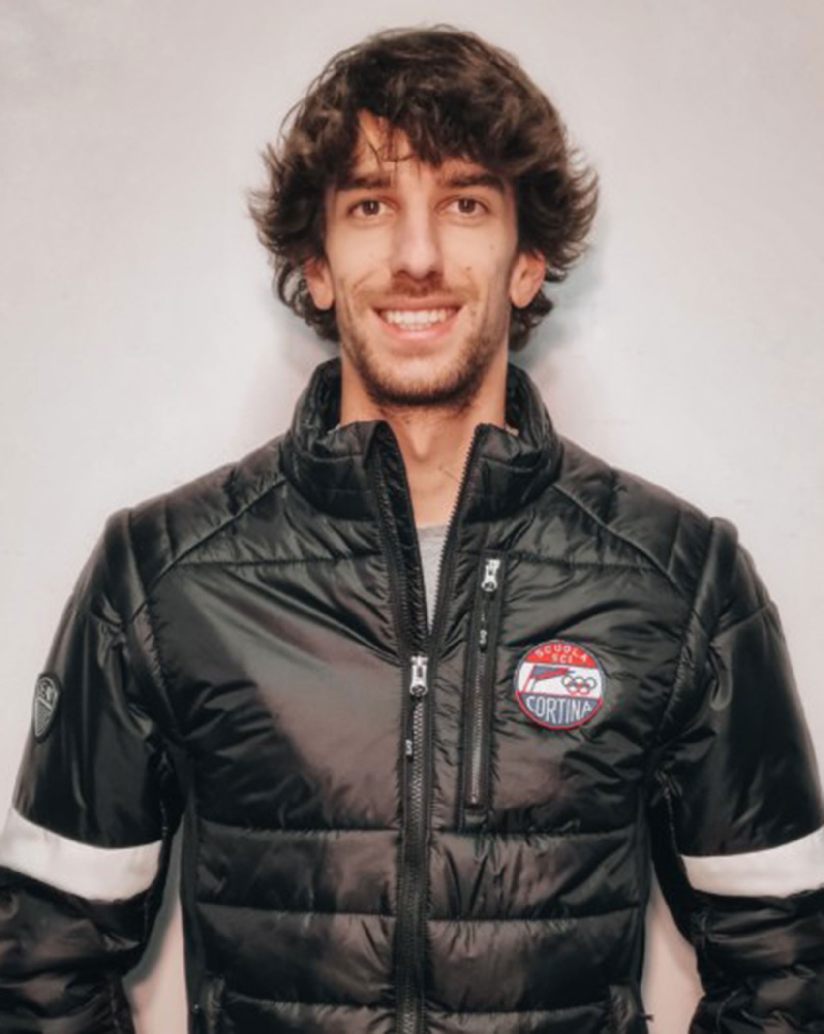 Man with wavy hair in a black Cortina jacket, smiling in front of a plain light-colored wall. - Scuola Sci Cortina