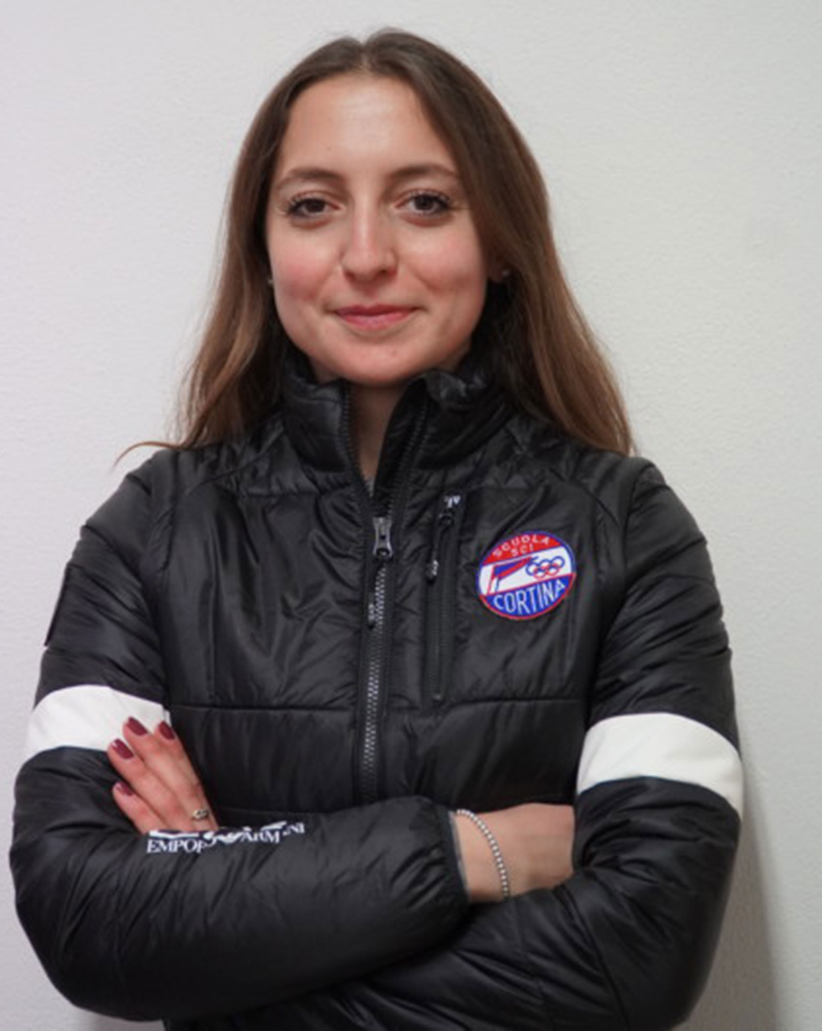 Woman in a black puffer jacket with arms crossed, standing against a plain white background. - Scuola Sci Cortina