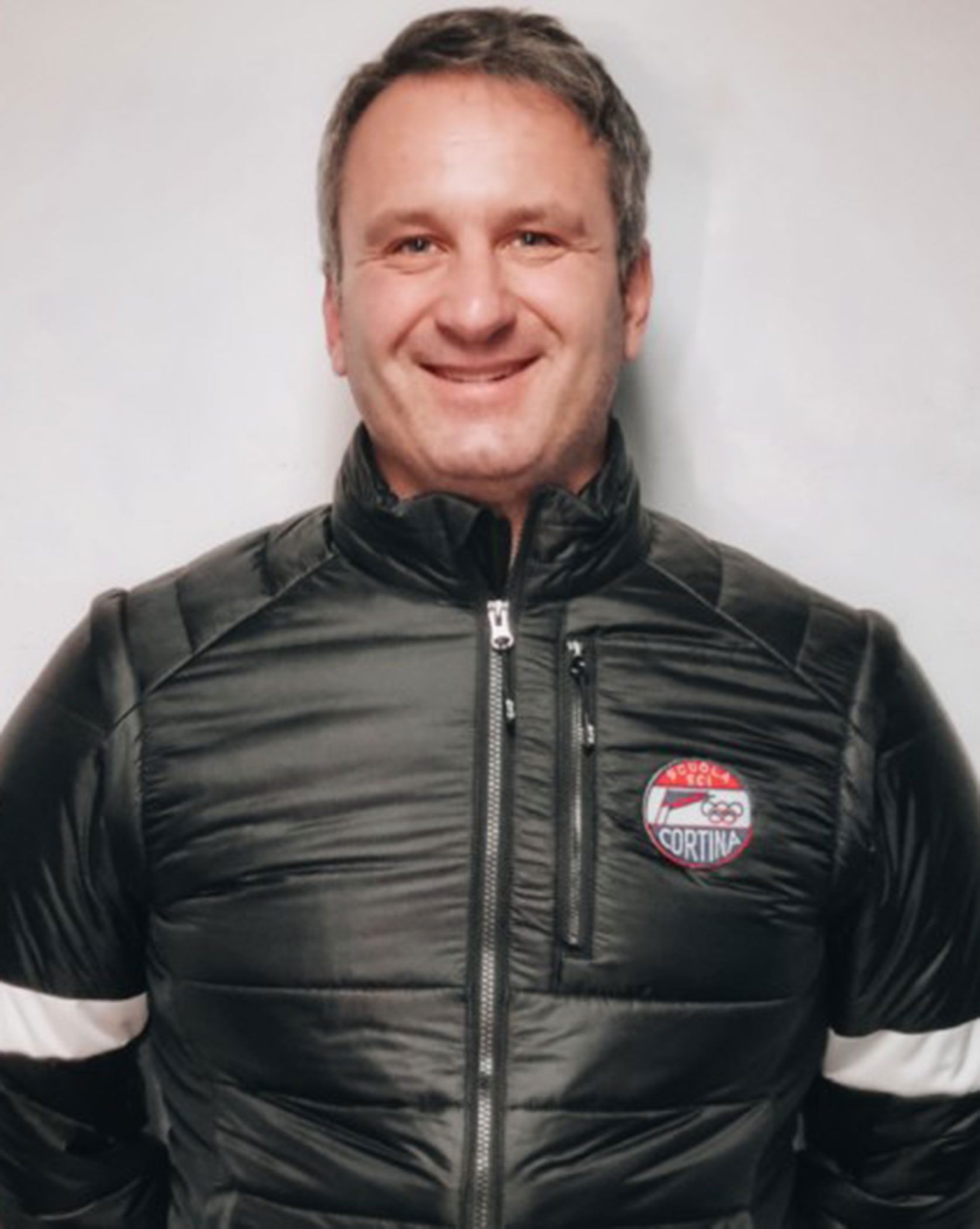 Man in a black jacket with a Cortina patch, standing and smiling in front of a light background. - Scuola Sci Cortina
