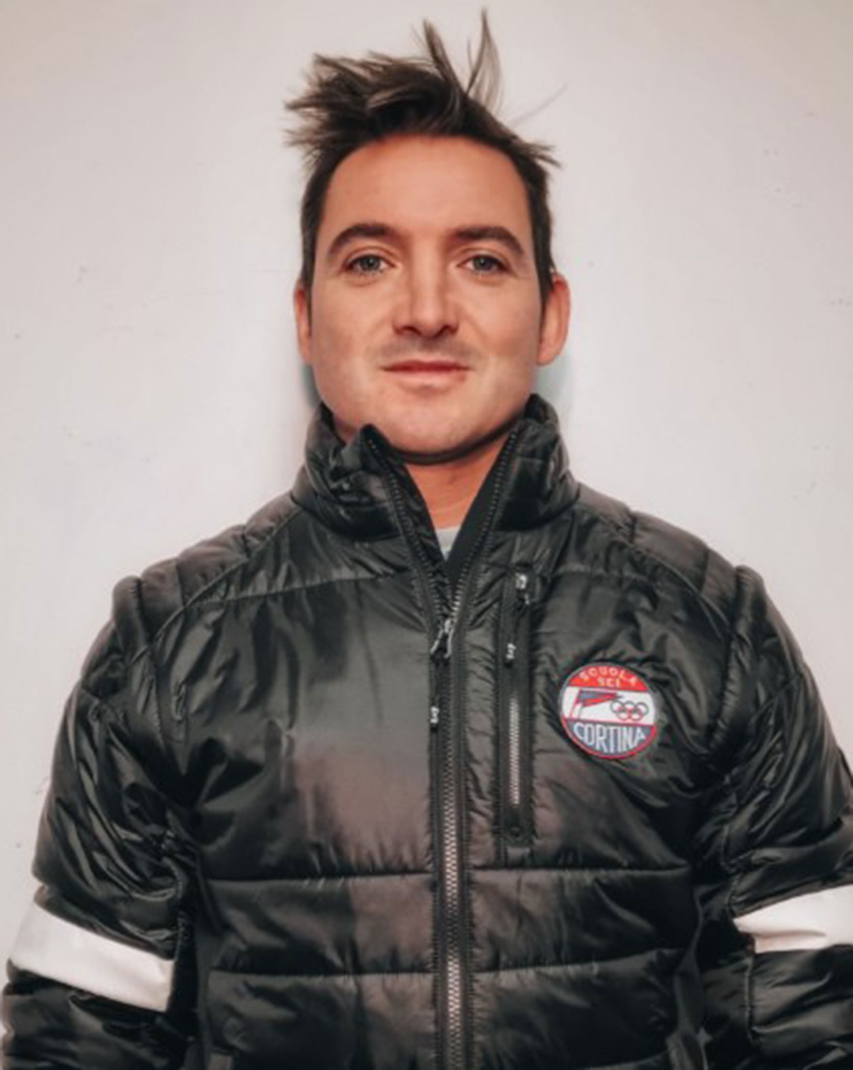 A person with short messy hair wears a black puffer jacket with a Cortina patch, standing against a plain wall. - Scuola Sci Cortina