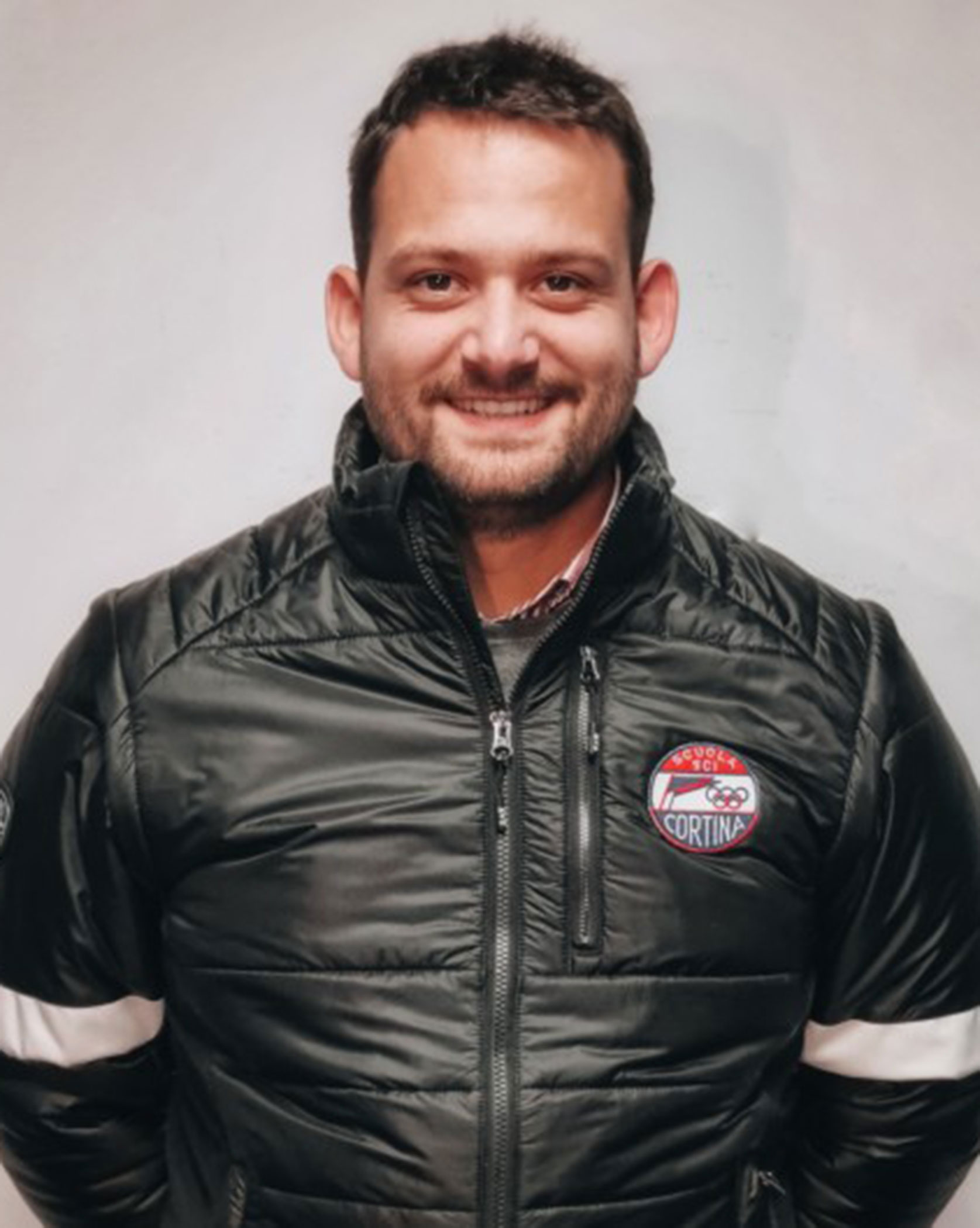 Man smiling, wearing a black puffer jacket with Cortina patch, standing against a plain background. - Scuola Sci Cortina
