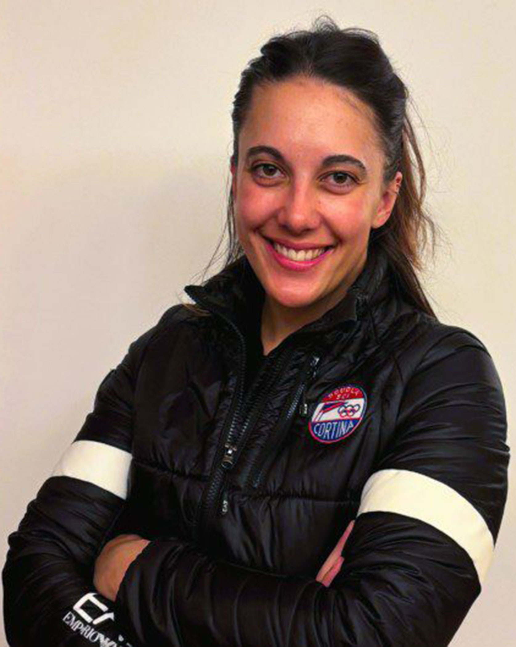 Woman with dark hair in a black jacket smiling, arms crossed, standing against a plain light background. - Scuola Sci Cortina