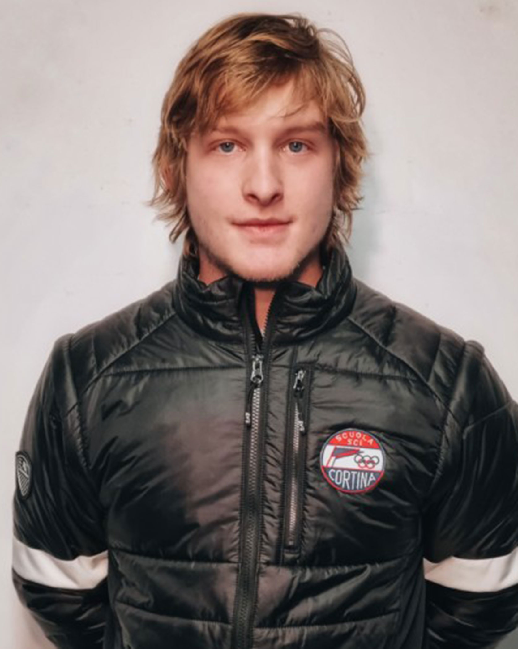 Young man with medium-length blonde hair wearing a black puffer jacket with a Cortina patch. - Scuola Sci Cortina