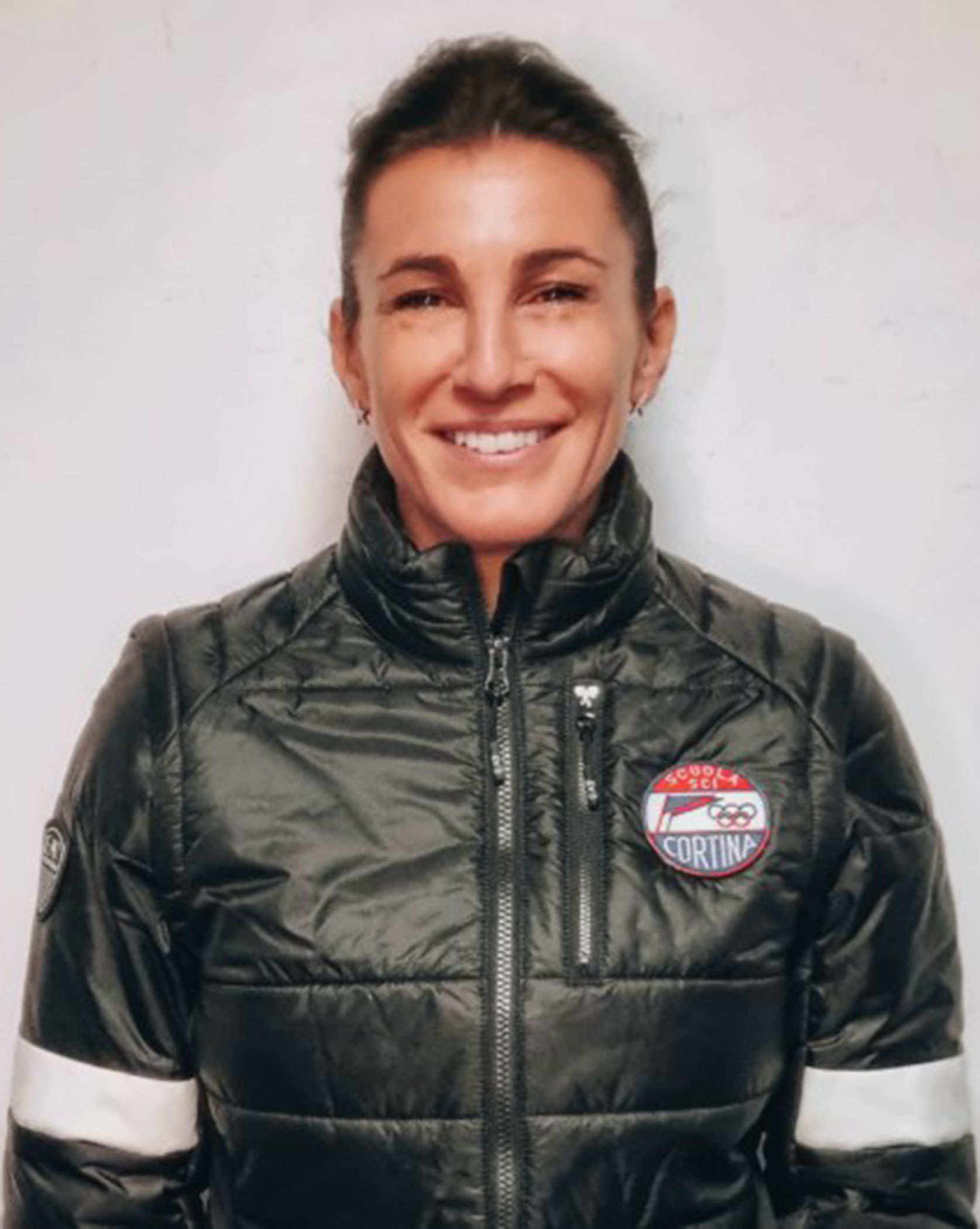 Smiling person in a puffy jacket with a Cortina patch, standing against a plain white background. - Scuola Sci Cortina