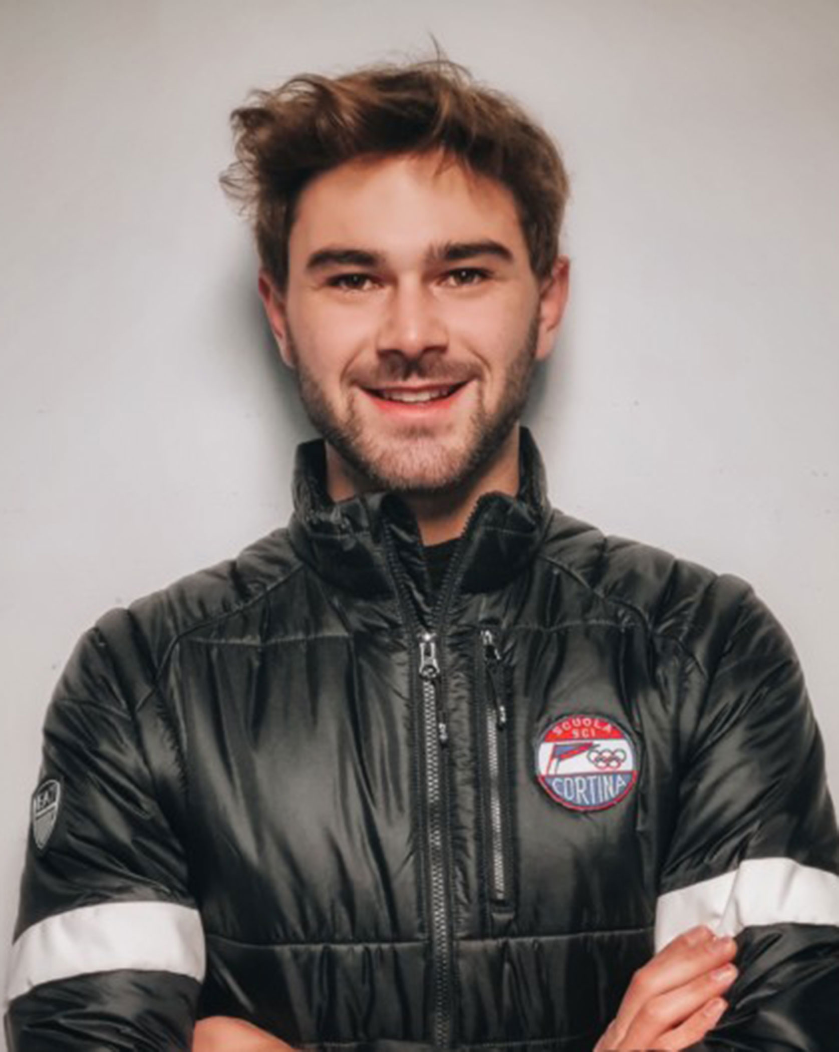 Man in a black puffer jacket with arms crossed, smiling in front of a plain light-colored wall. - Scuola Sci Cortina