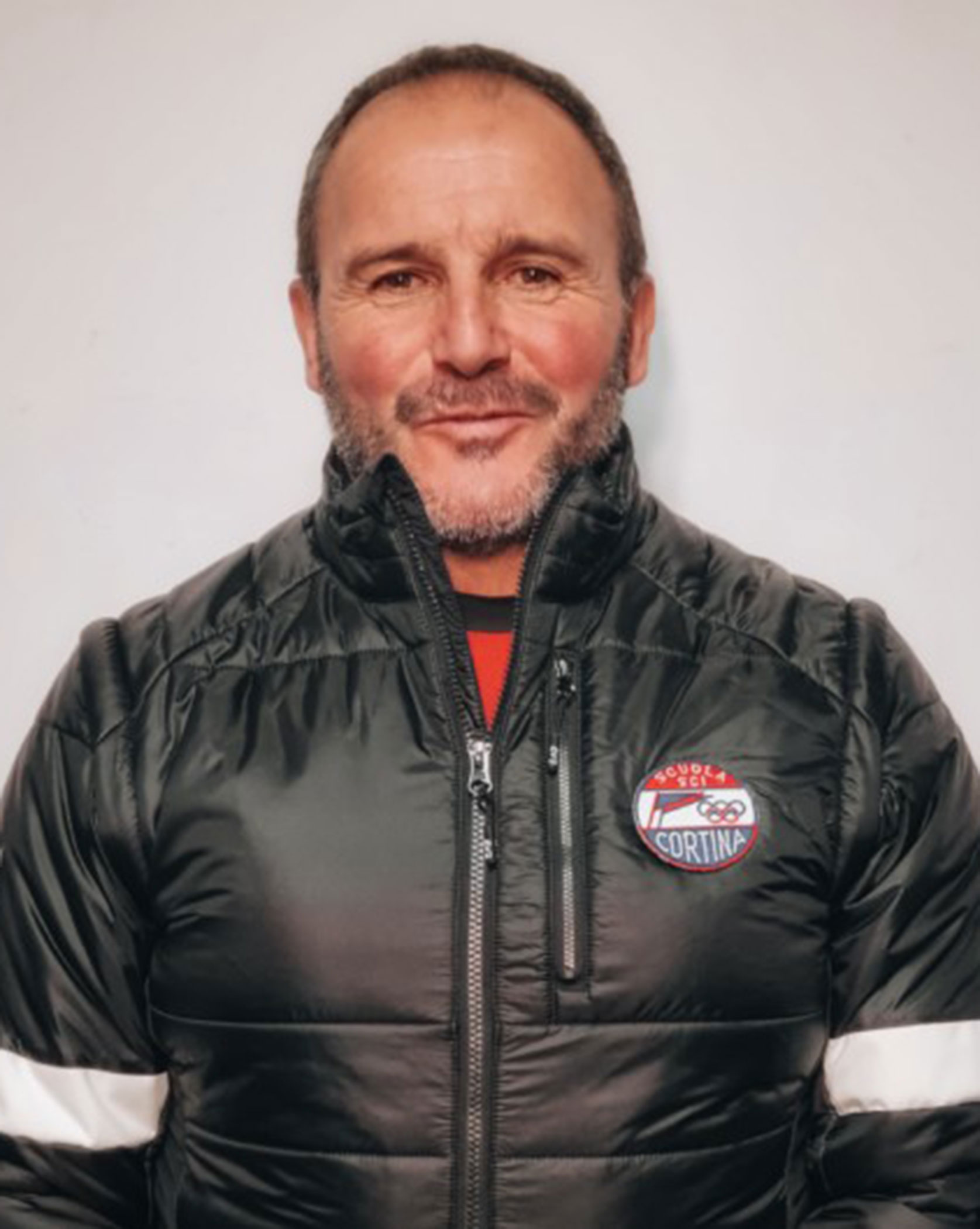 Man in a black puffer jacket with a badge, standing against a plain light background, smiling slightly. - Scuola Sci Cortina
