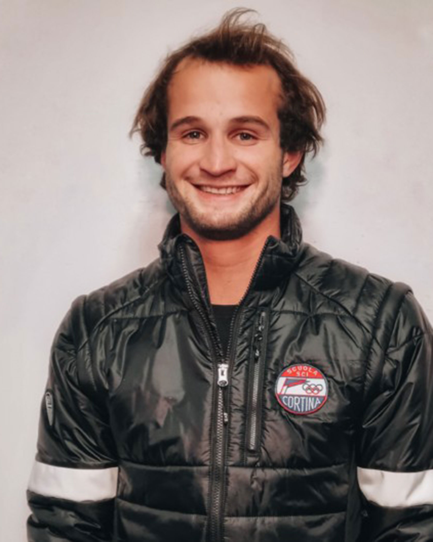 Smiling person wearing a black puffy jacket with a Cortina logo, standing against a light background. - Scuola Sci Cortina