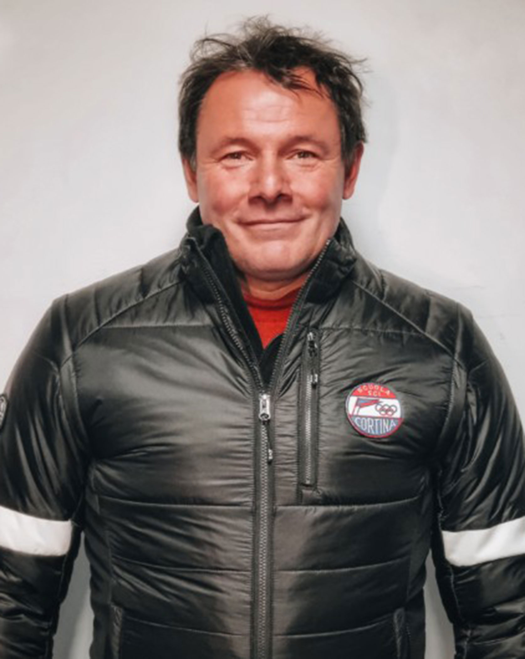 Smiling man in a black winter jacket with a Cortina patch, standing against a plain light background. - Scuola Sci Cortina