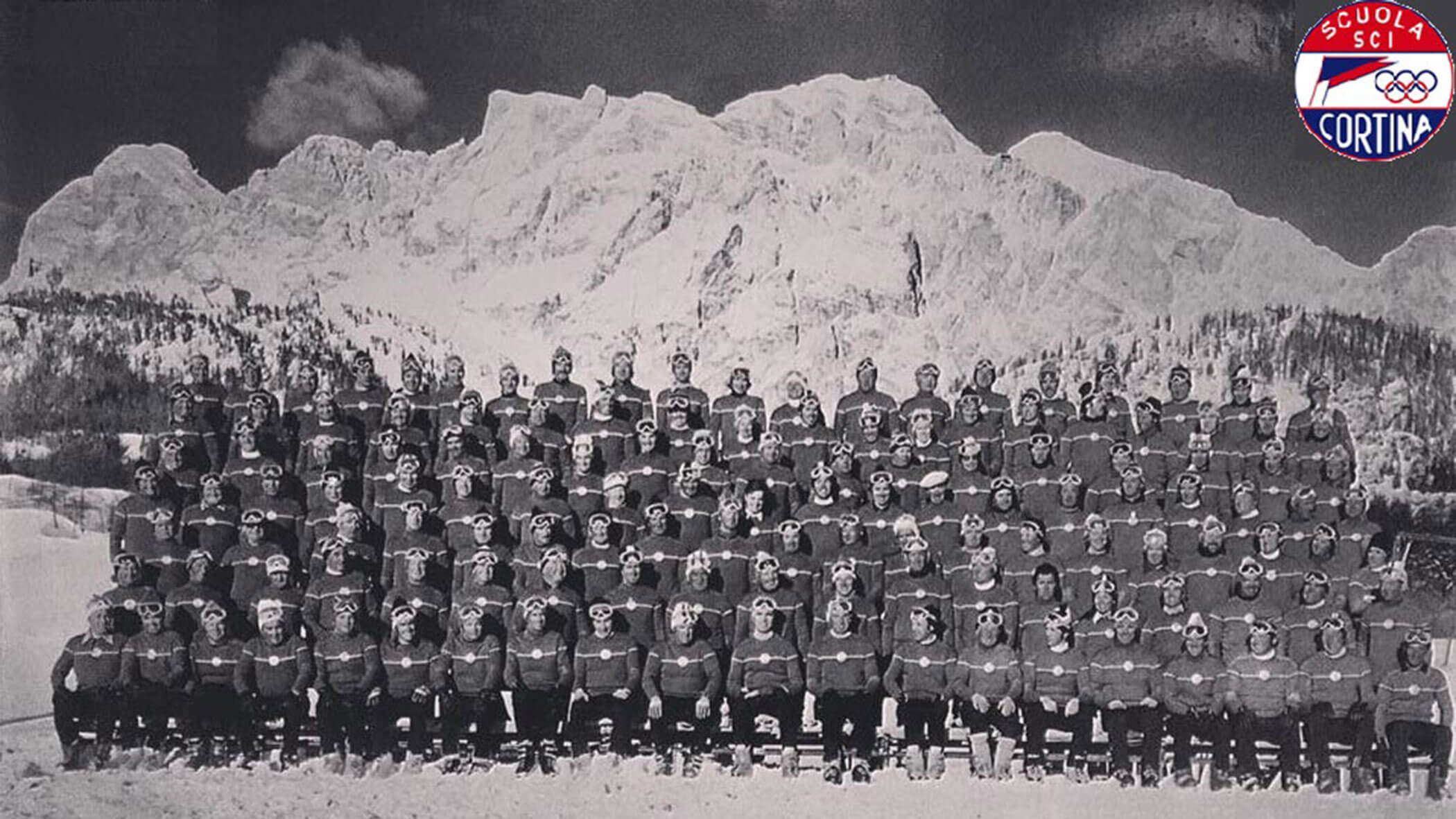 Large group of people in winter gear pose before snowy mountains; Scuola Sci Cortina logo in corner. - Scuola Sci Cortina