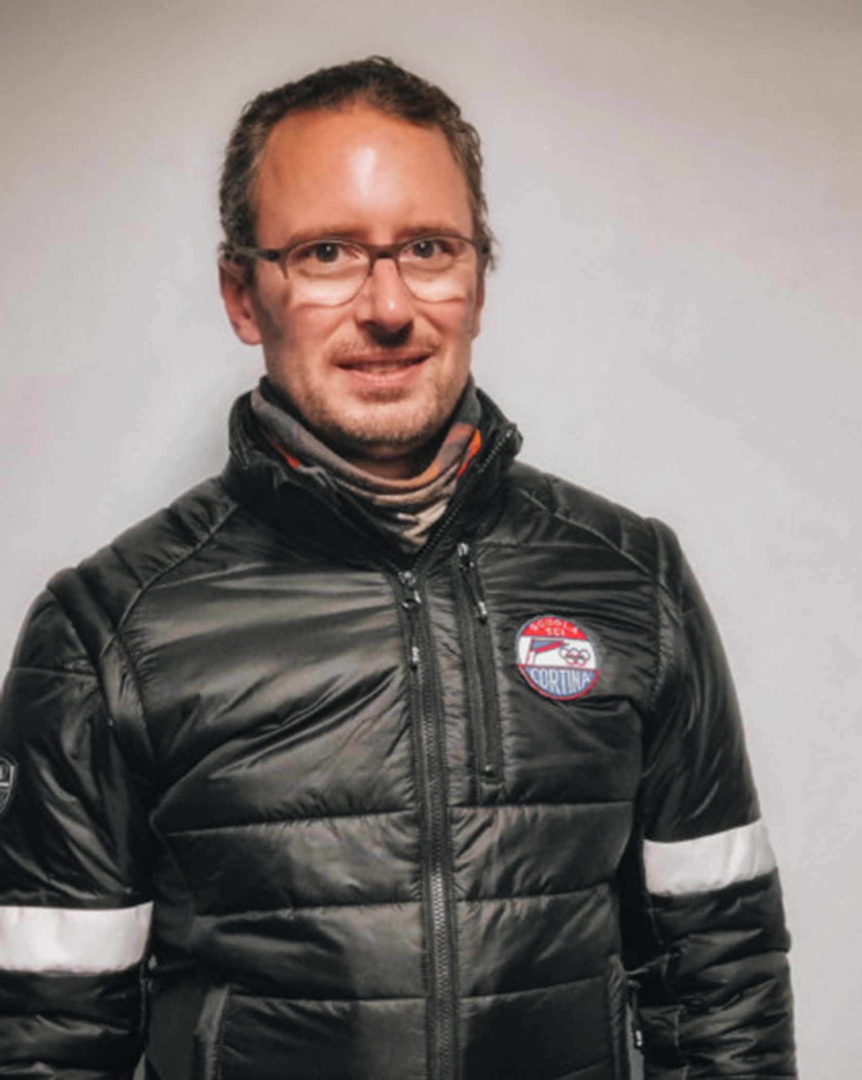 A person in a black jacket with a Cortina badge stands against a plain light-colored background. - Scuola Sci Cortina
