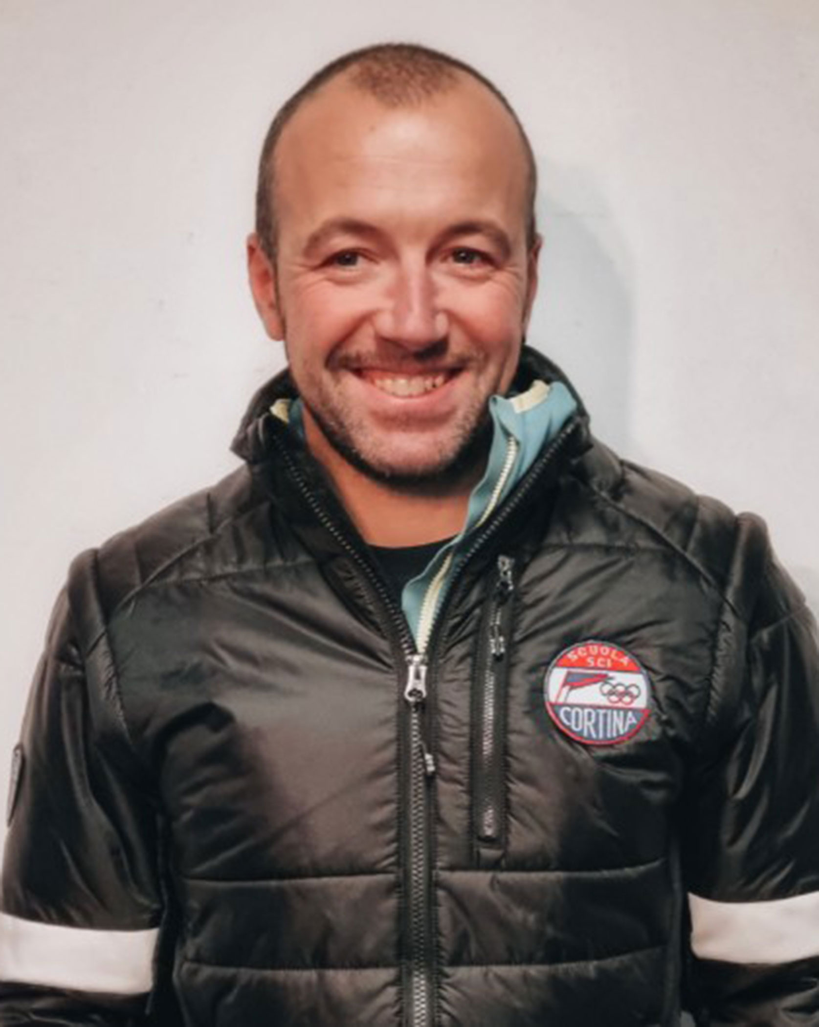 Smiling man in a black padded jacket with a Cortina badge, standing against a plain light background. - Scuola Sci Cortina