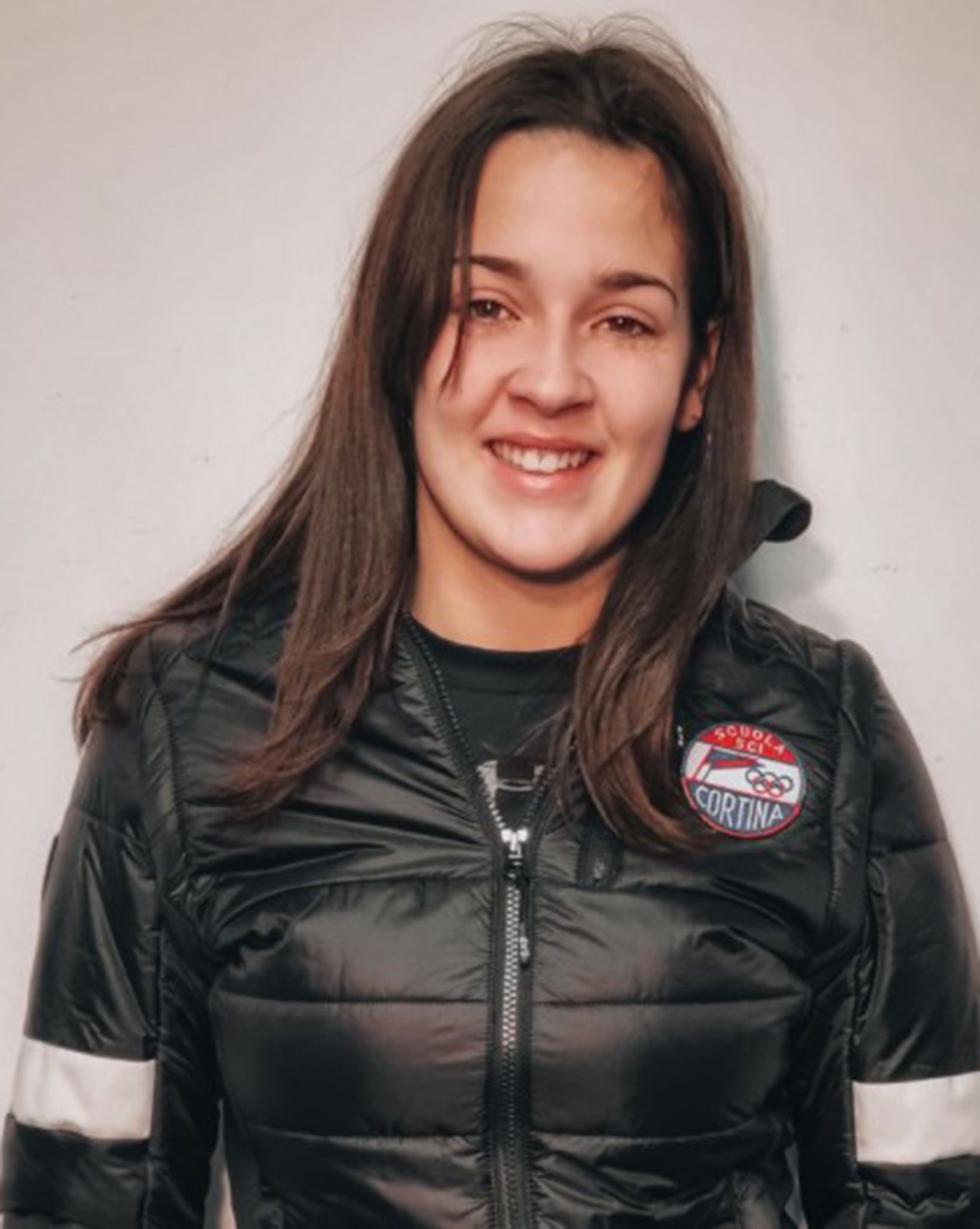Woman with straight brown hair in a black Cortina jacket, smiling at the camera against a plain background. - Scuola Sci Cortina