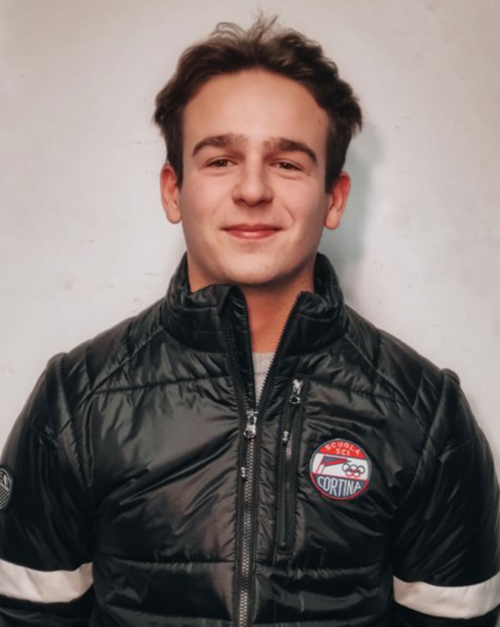 Young man in a black puffer jacket with a Cortina badge, smiling against a plain background. - Scuola Sci Cortina