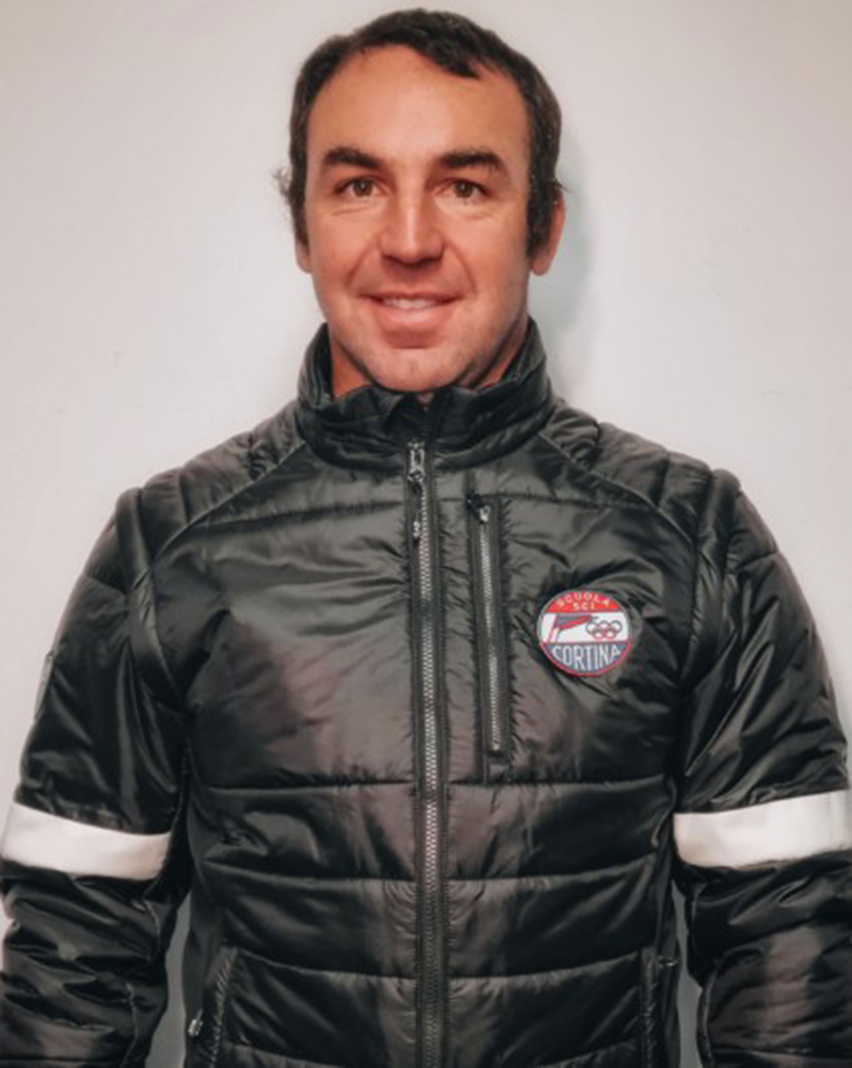 Man in a black puffer jacket with a Cortina badge, standing against a plain light background, smiling. - Scuola Sci Cortina