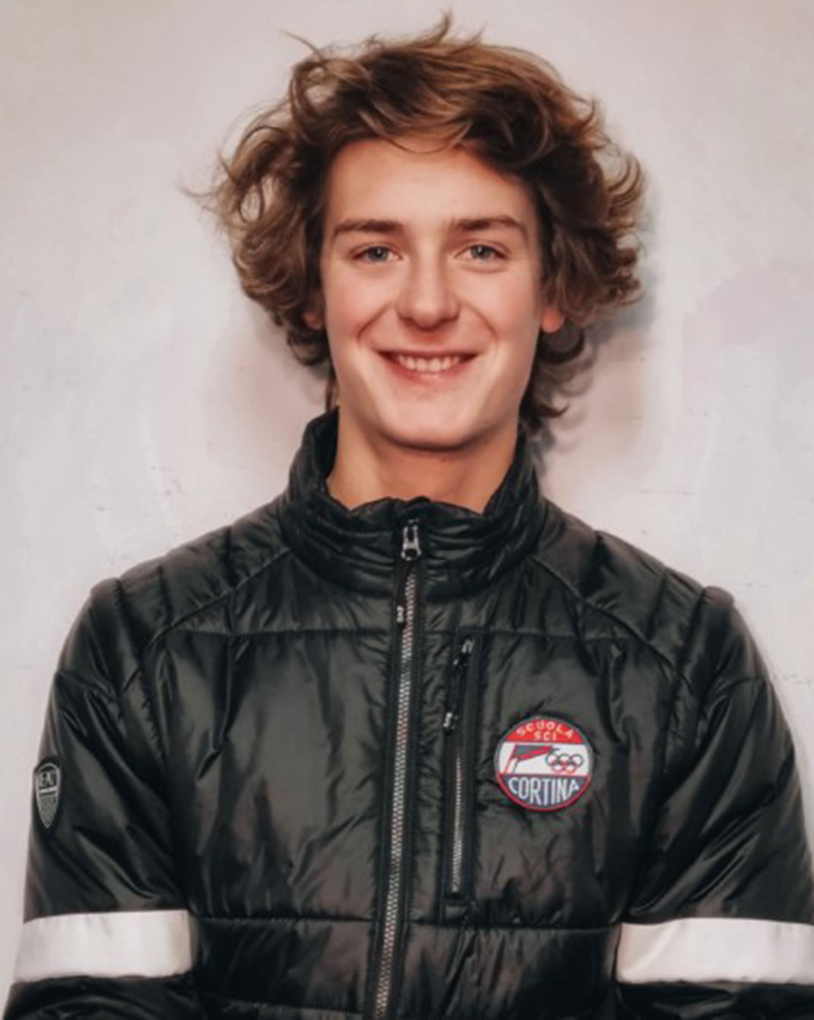 Young person with wavy brown hair, smiling, wearing a black jacket with a red and white Cortina logo patch. - Scuola Sci Cortina