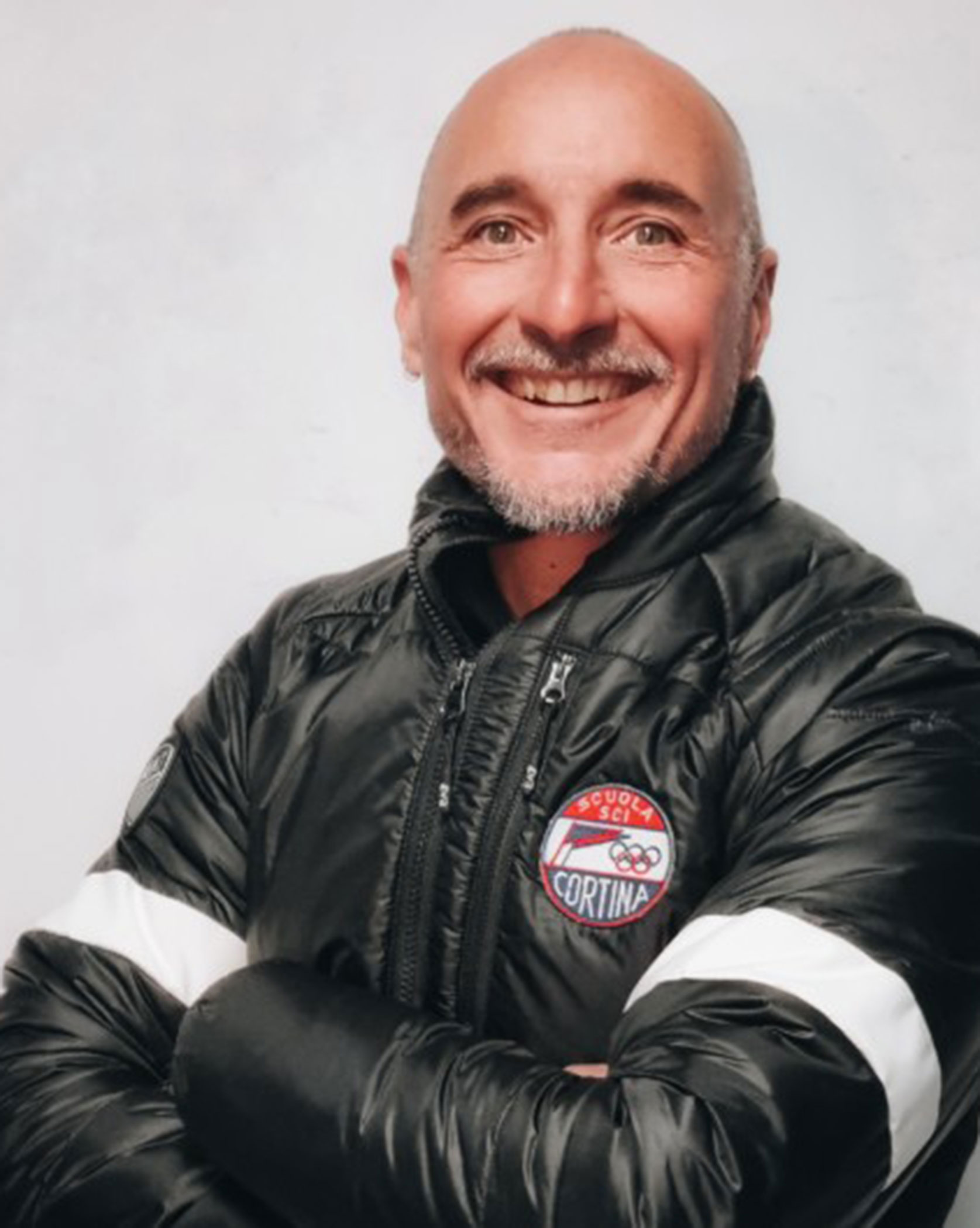 Smiling person in a black puffer jacket with a Cortina logo, arms crossed, against a plain background. - Scuola Sci Cortina