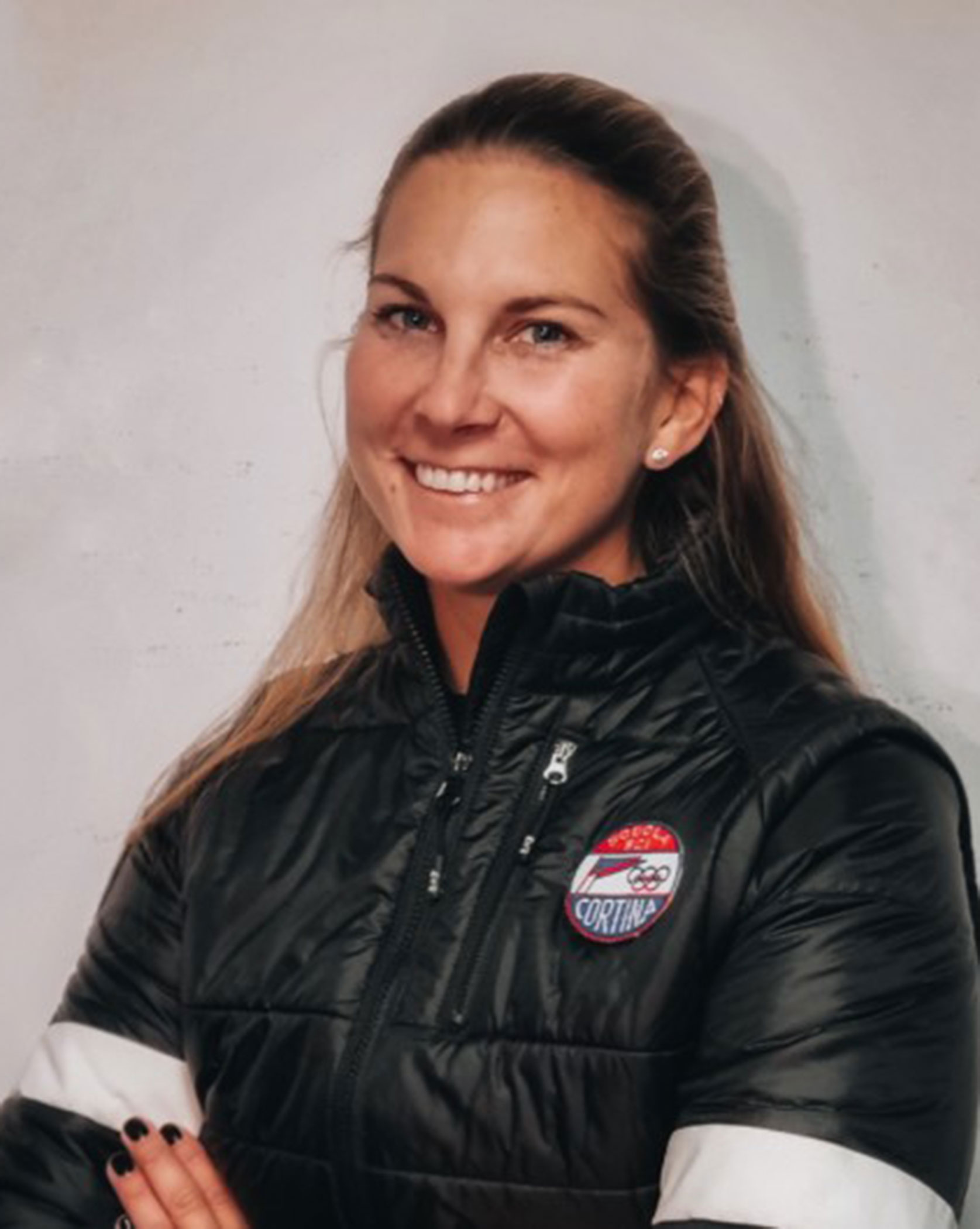 Woman in a black jacket with a Cortina patch, smiling with arms crossed against a plain background. - Scuola Sci Cortina