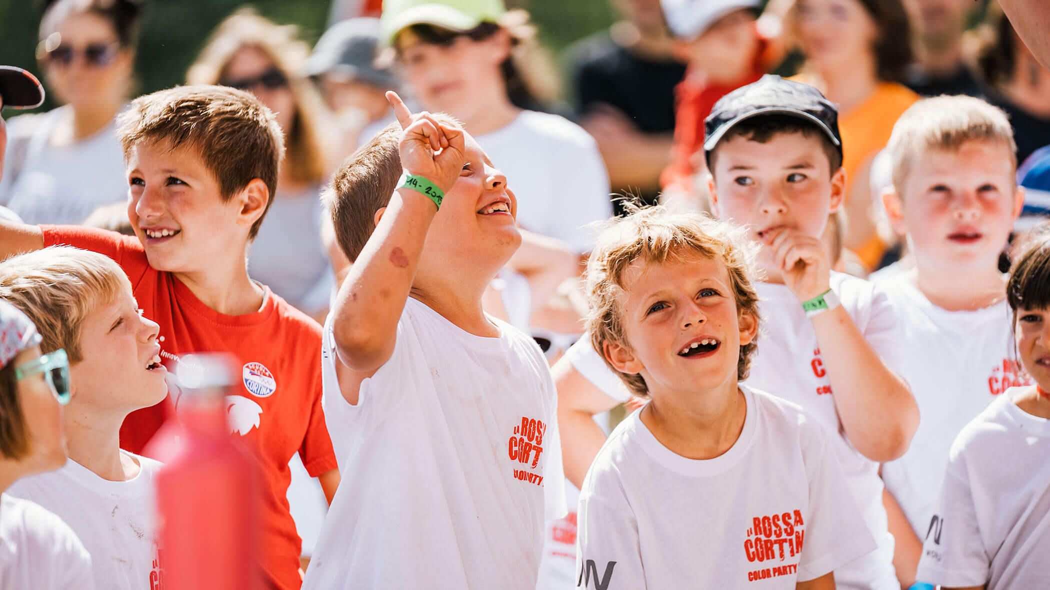 Children smiling and looking up, wearing white shirts, outdoors in a lively, sunny group setting. - Scuola Sci Cortina