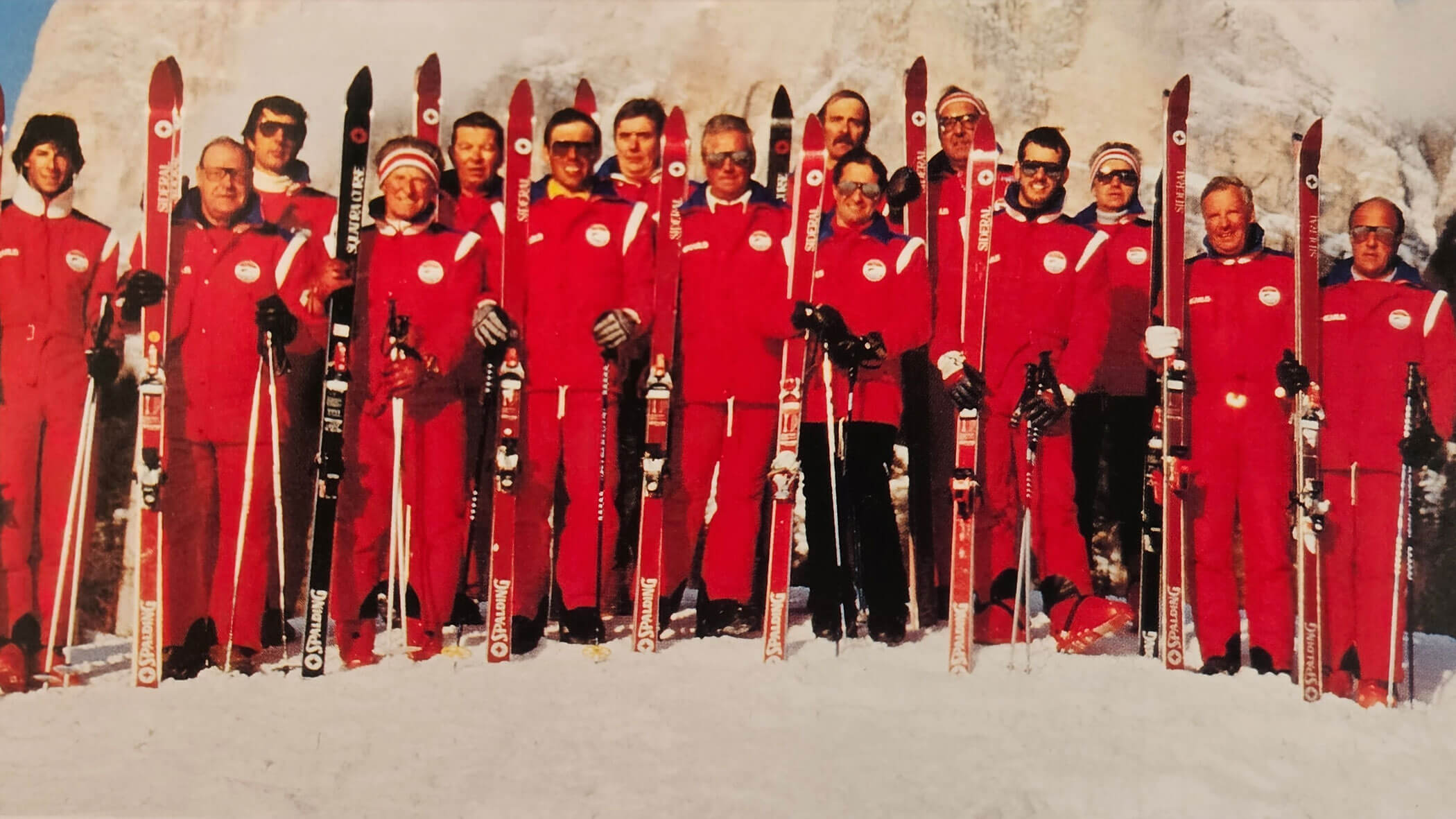 A group of people in matching red ski suits stand in the snow holding skis side by side. - Scuola Sci Cortina