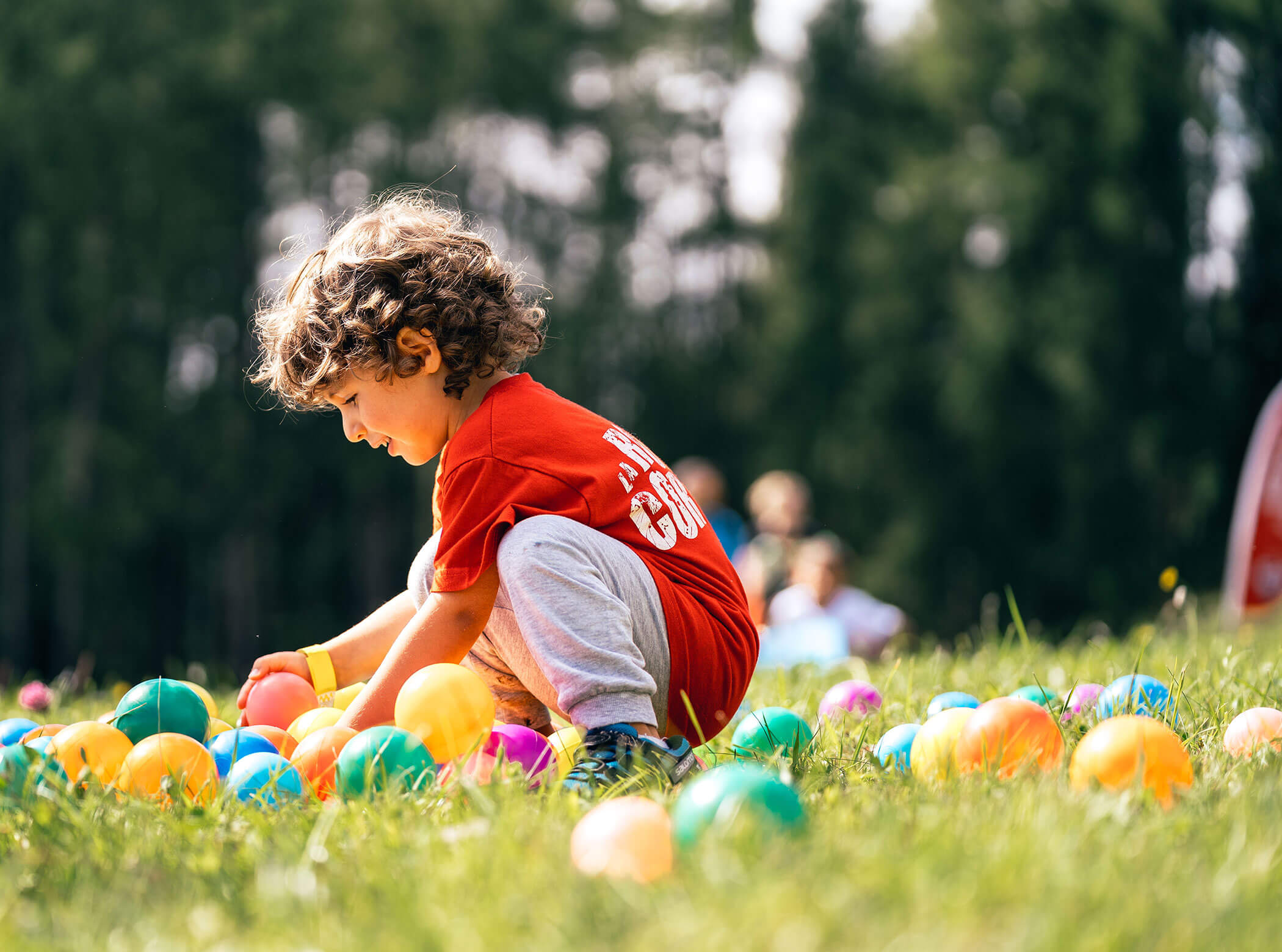 A young child in a red shirt plays with colorful plastic balls on green grass outdoors. - Scuola Sci Cortina