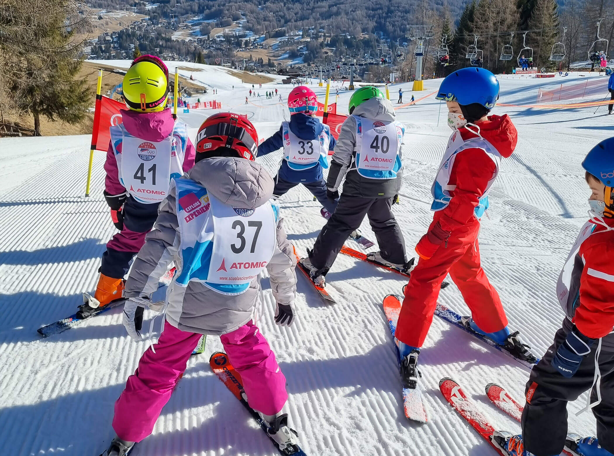 Children in ski gear stand in a group on a snowy slope, preparing to ski with numbered vests on. - Scuola Sci Cortina