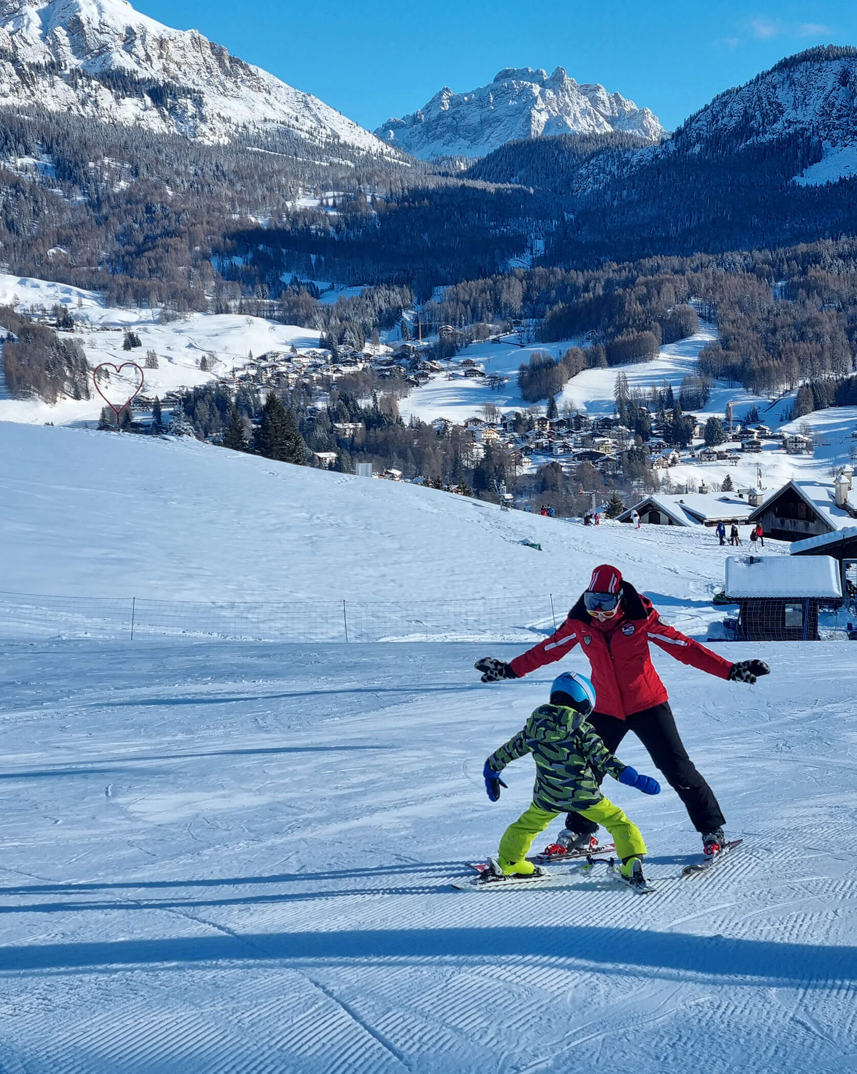 Adult teaching child to ski on a snowy mountain slope with village and mountains in the background. - Scuola Sci Cortina