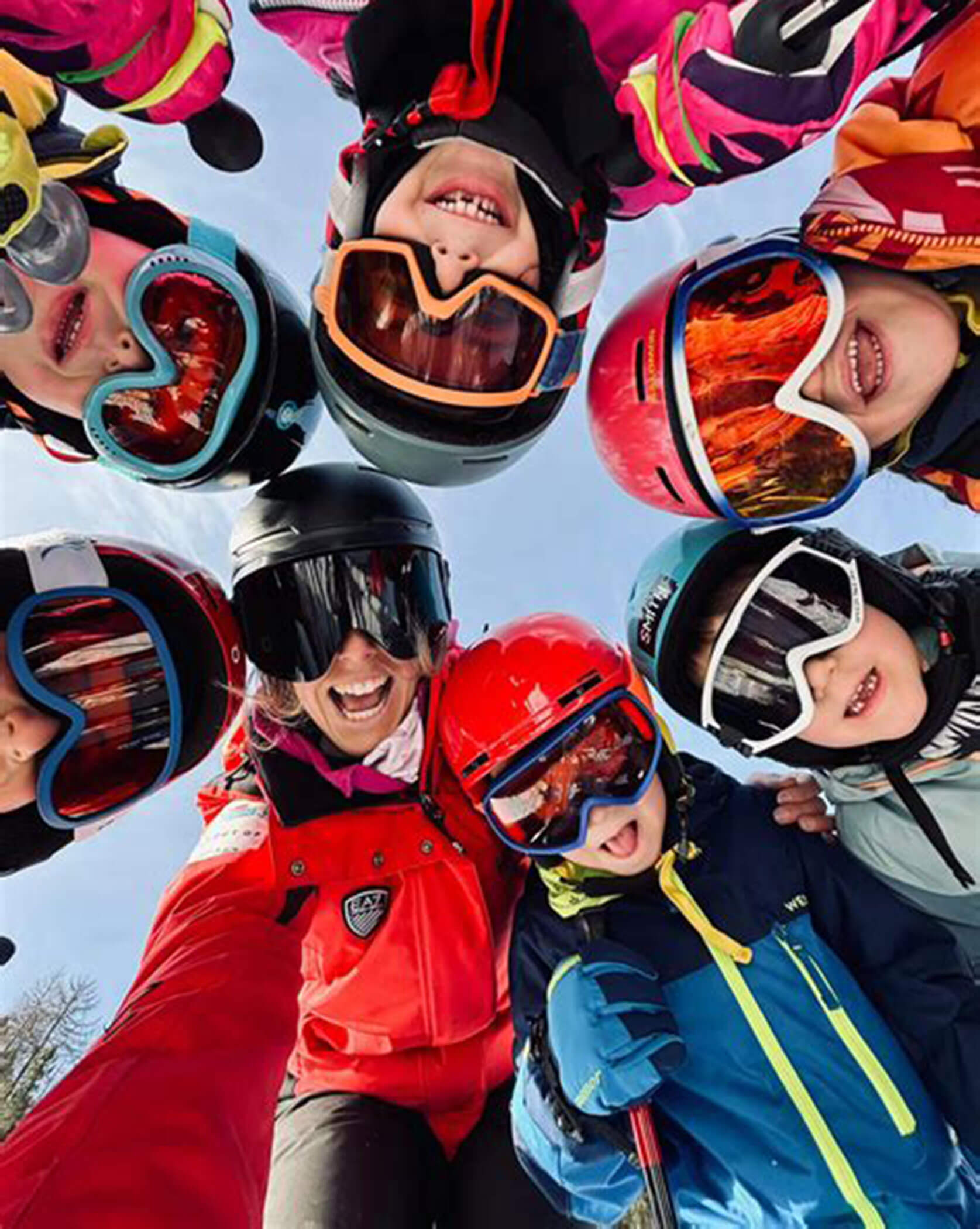 Group of kids and an adult in ski gear huddled together, smiling up at the camera on a snowy day. - Scuola Sci Cortina