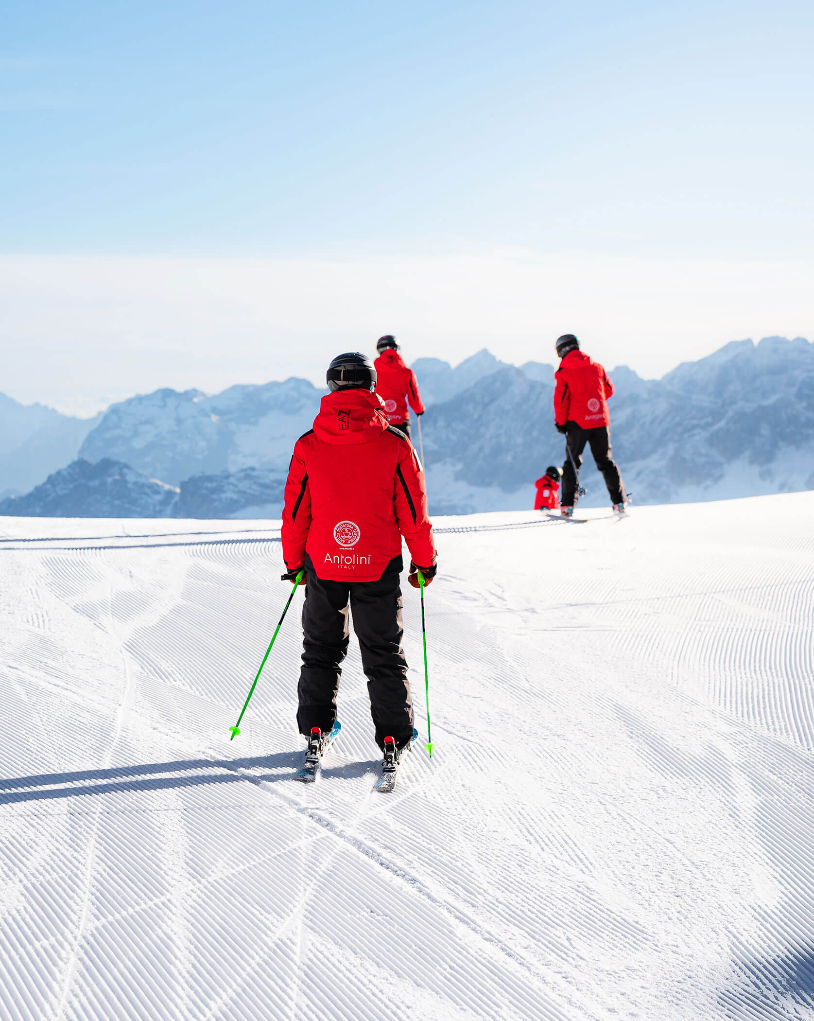 Three skiers in red jackets stand on a snowy slope with mountains in the background. - Scuola Sci Cortina