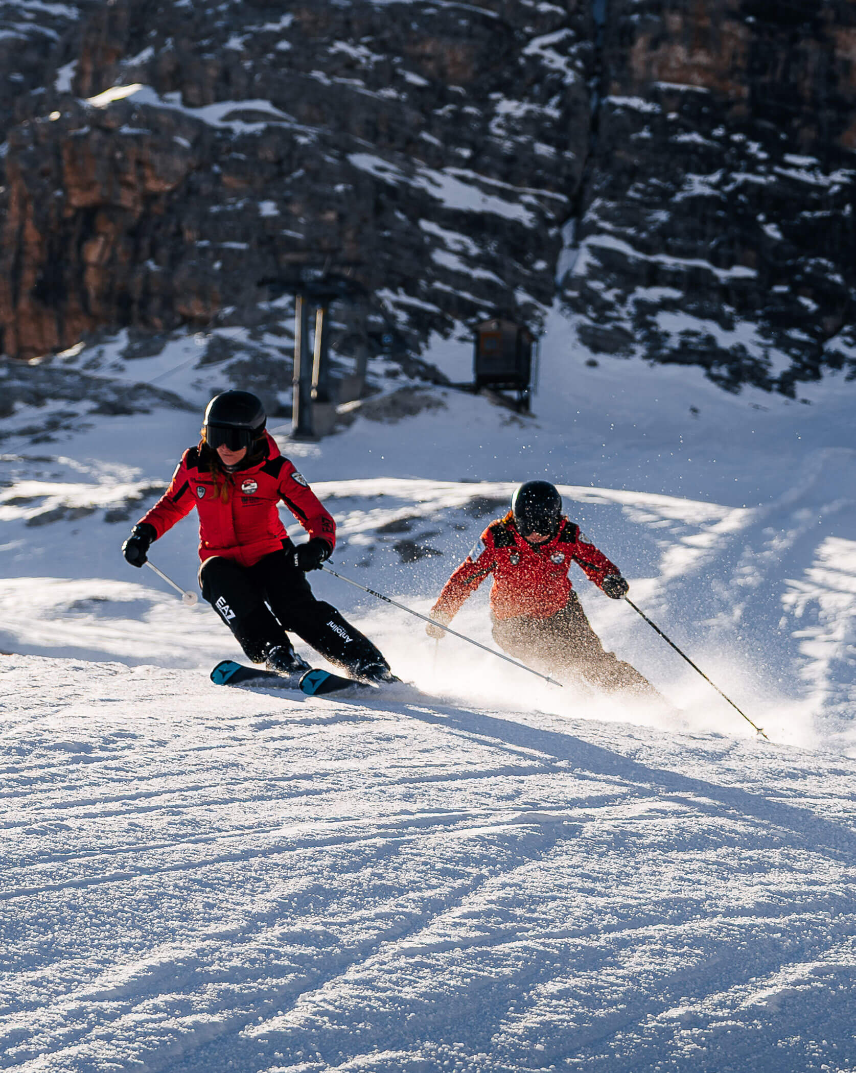 Two skiers in red jackets ski downhill on a snowy slope with rocky cliffs in the background. - Scuola Sci Cortina