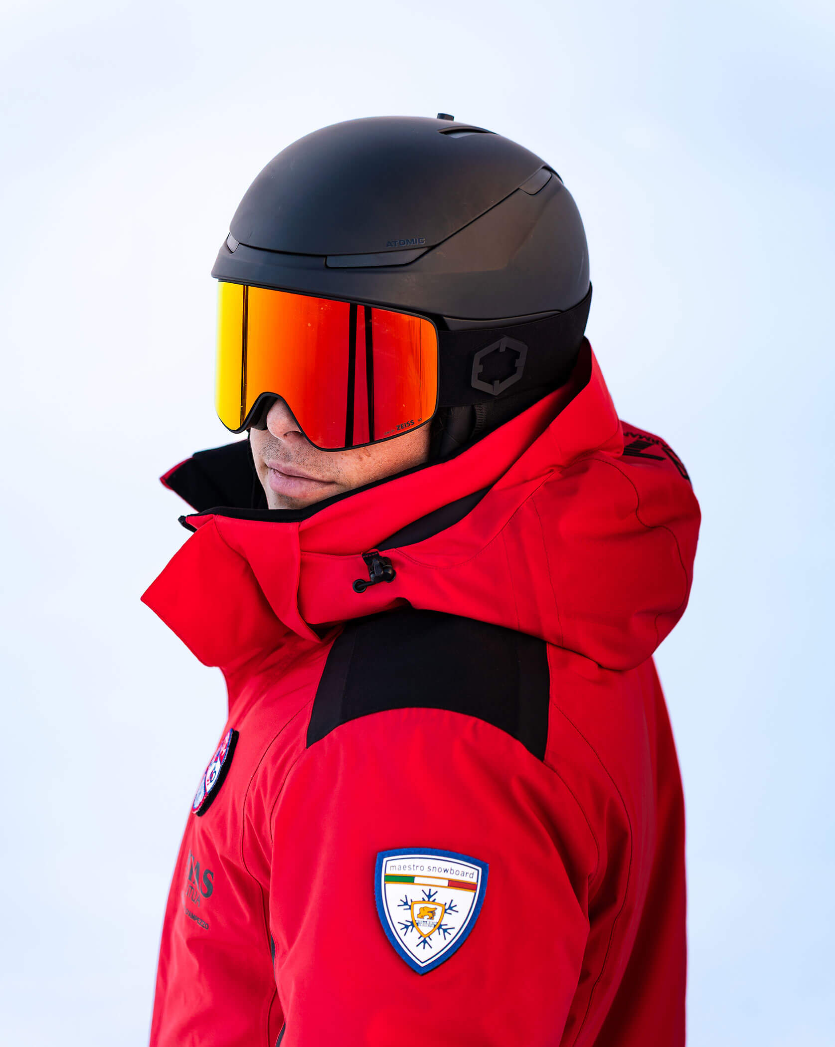 Person in red jacket, ski helmet, and orange goggles stands against a snowy background, facing left. - Scuola Sci Cortina