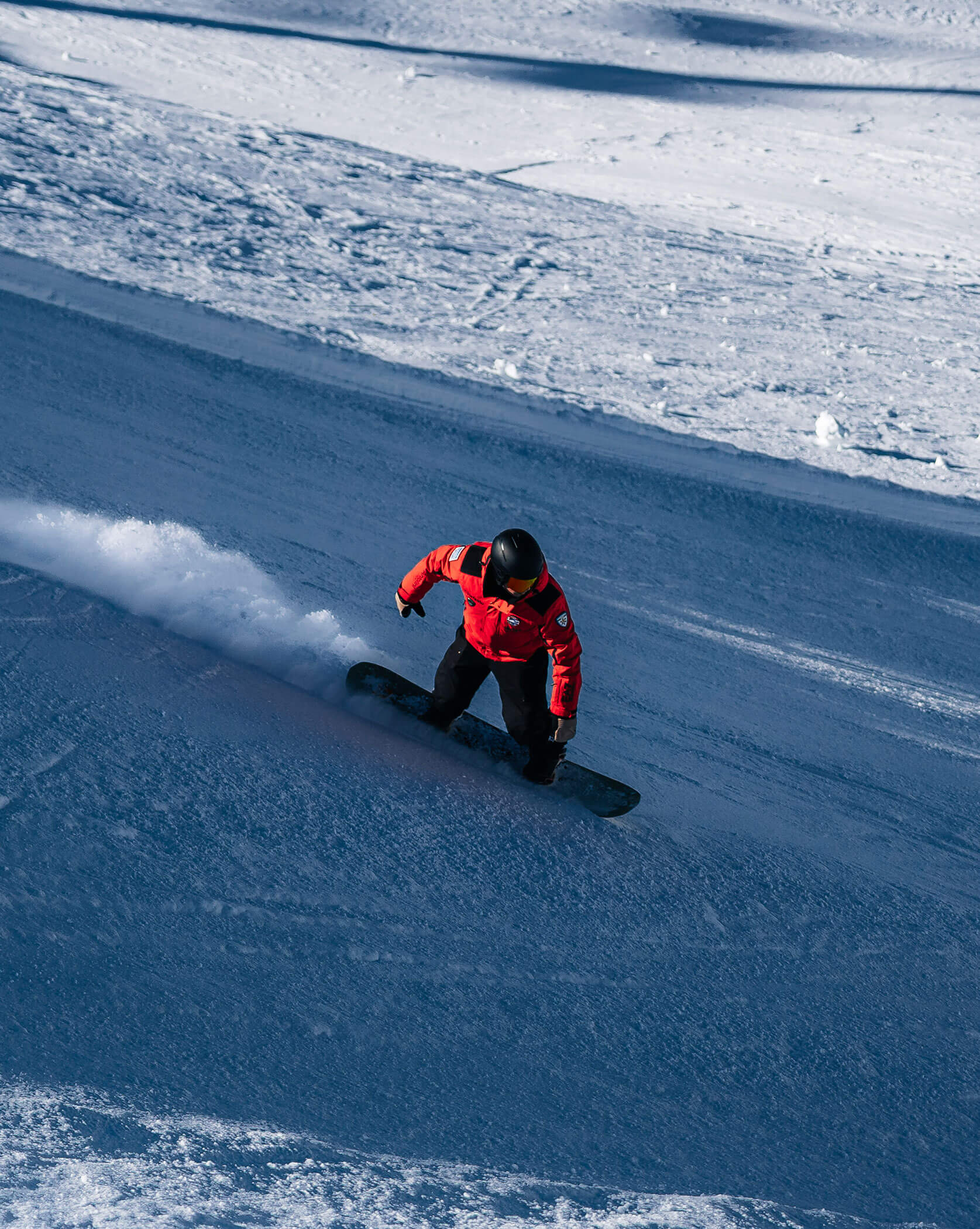 Snowboarder in red jacket riding down a snowy slope, making a turn and leaving a trail of snow behind. - Scuola Sci Cortina