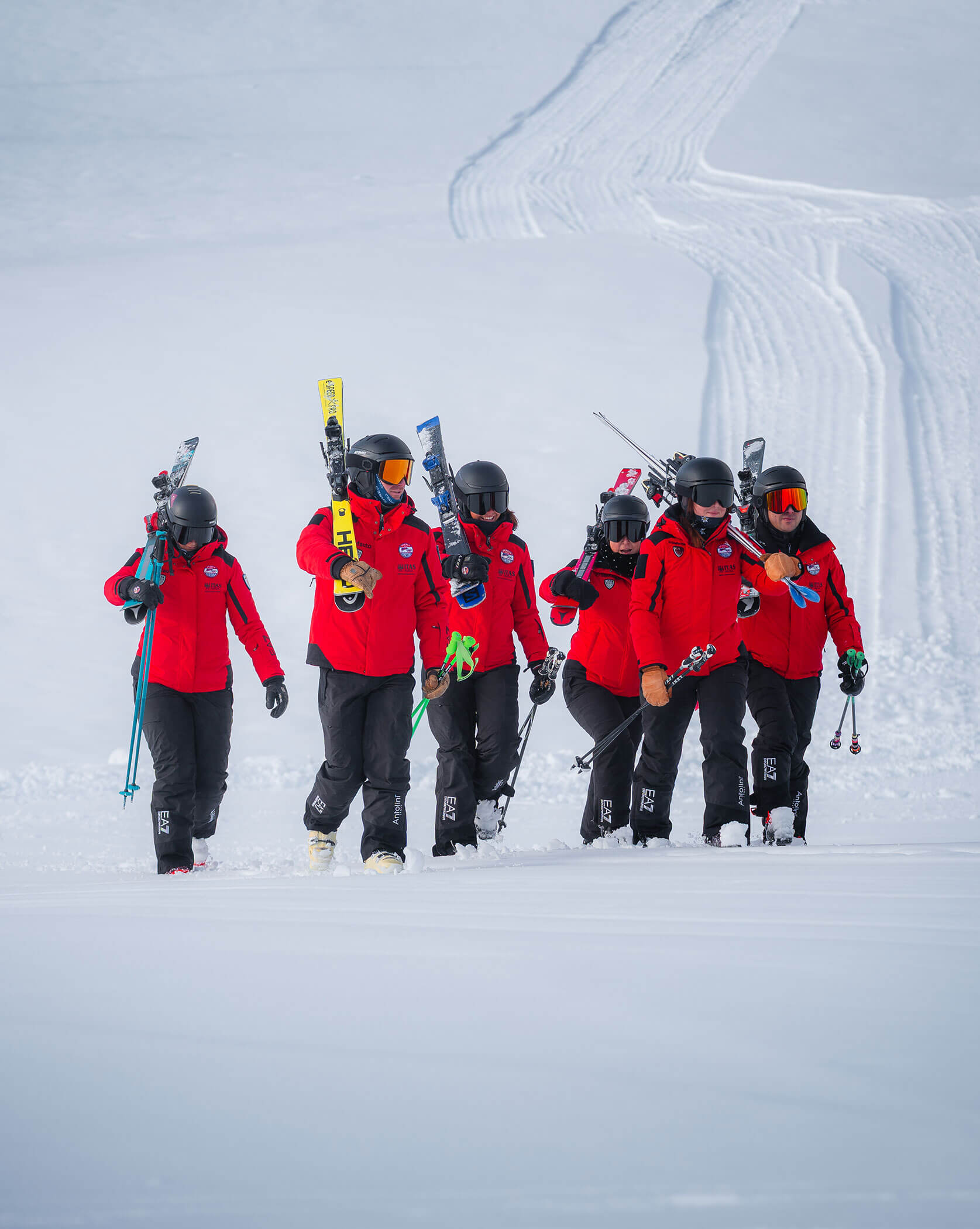 Six skiers in red jackets and helmets walk uphill on snow, carrying skis over their shoulders. - Scuola Sci Cortina